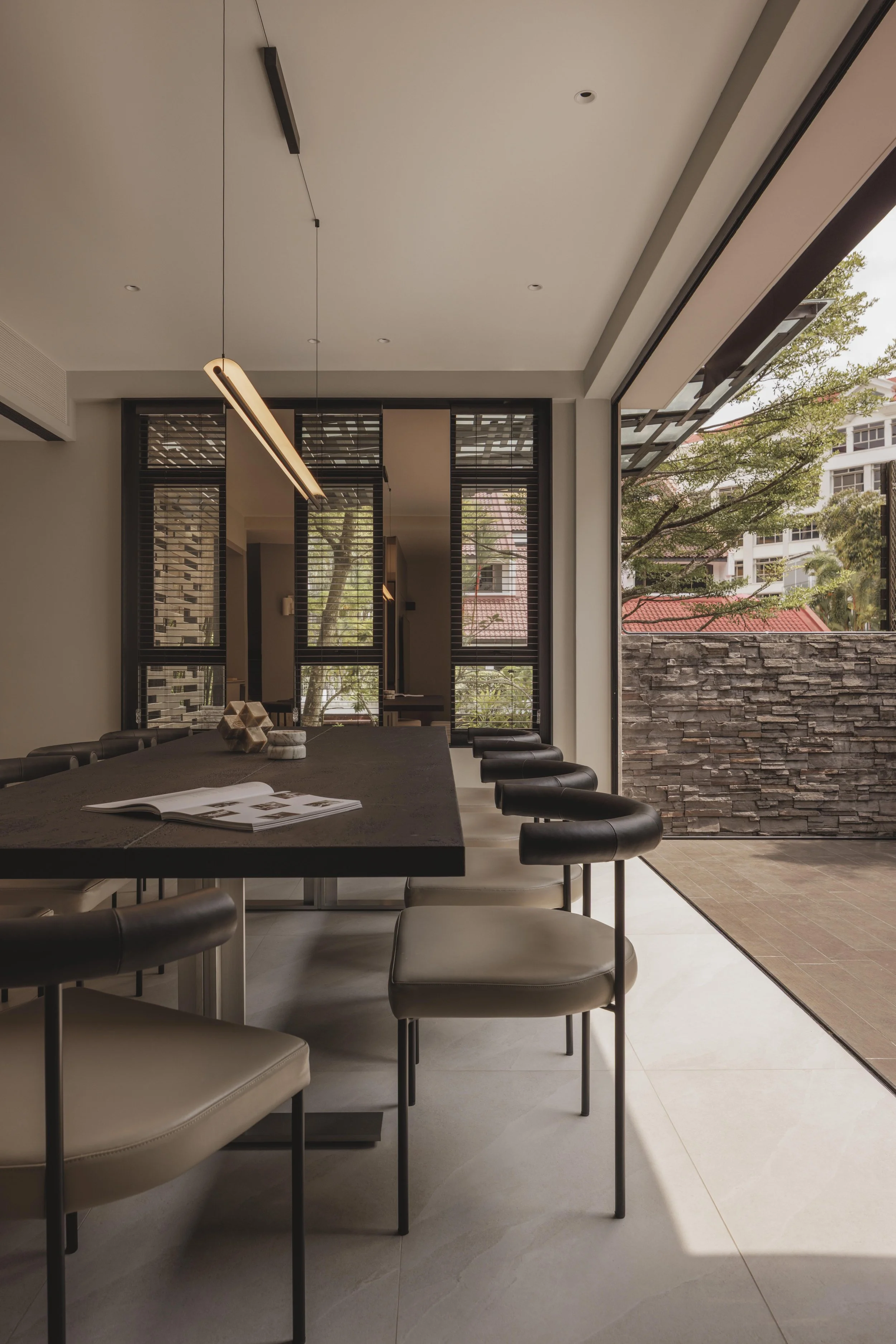 Modern dining room with a long black table surrounded by cream-colored chairs with black backs, open sliding glass doors revealing a backyard with trees and a stone fence, and a minimalist pendant light hanging from the ceiling.