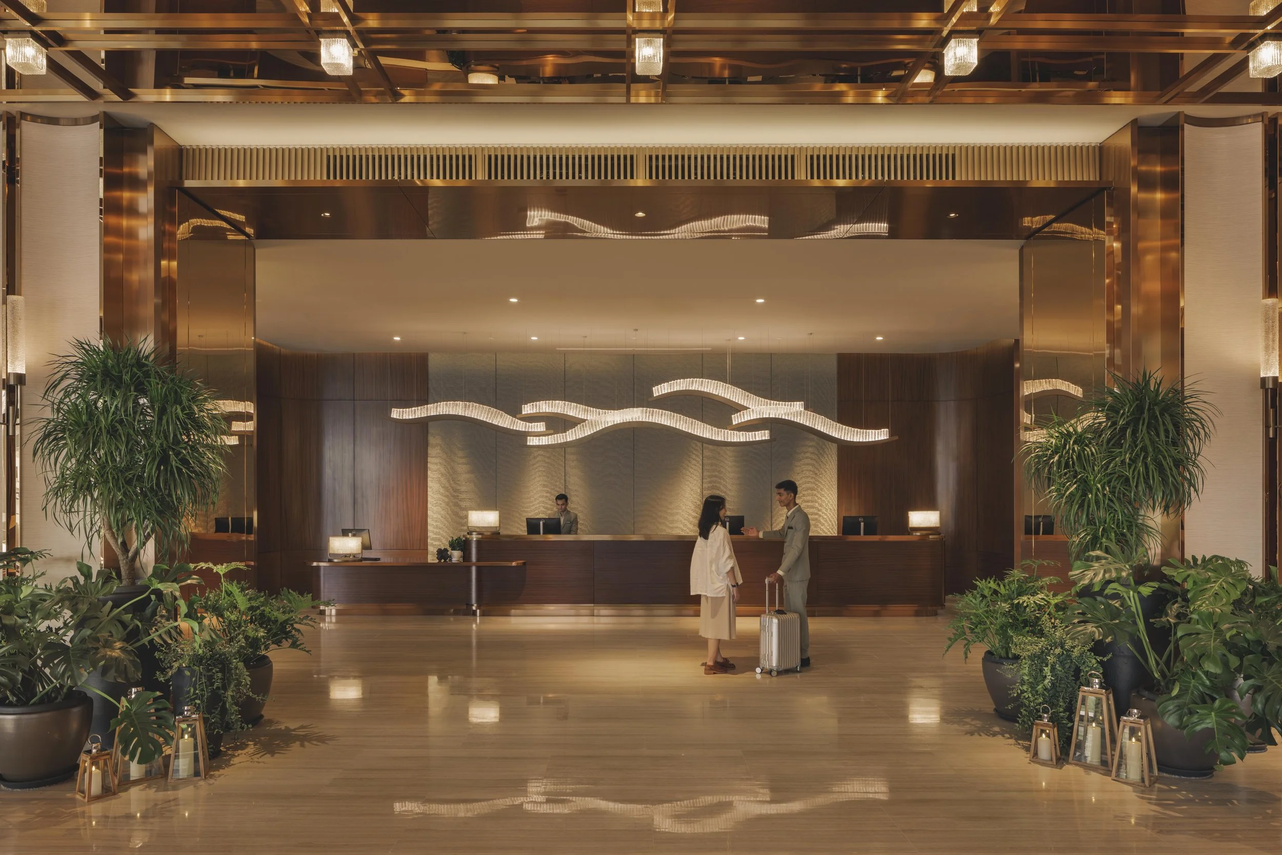 Hotel lobby reception area with two people at the counter, surrounded by large potted plants and decorative lanterns, with warm lighting and wooden accents.