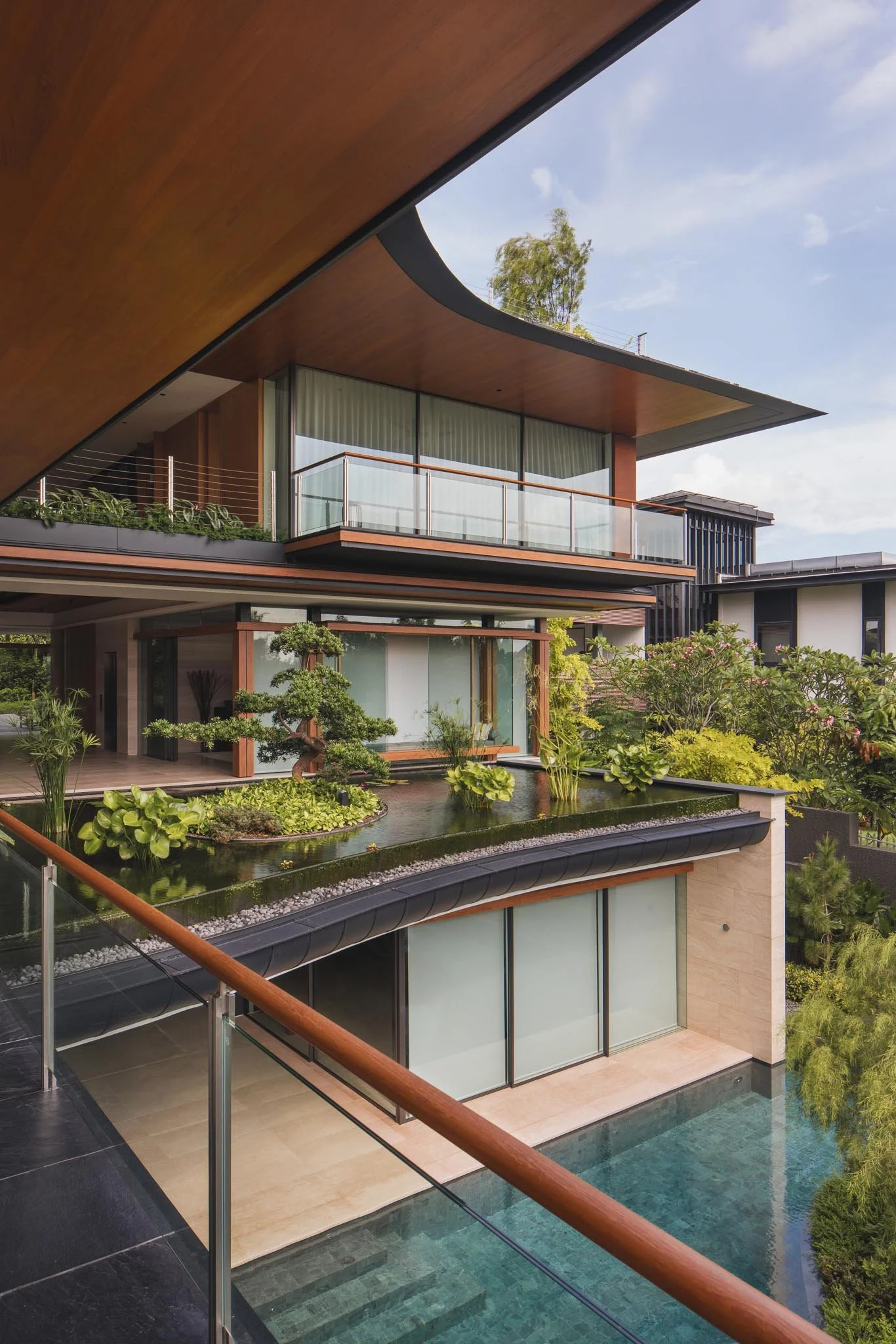 Modern multi-level house with large glass windows, garden, and swimming pool, seen from a balcony with wooden handrail.