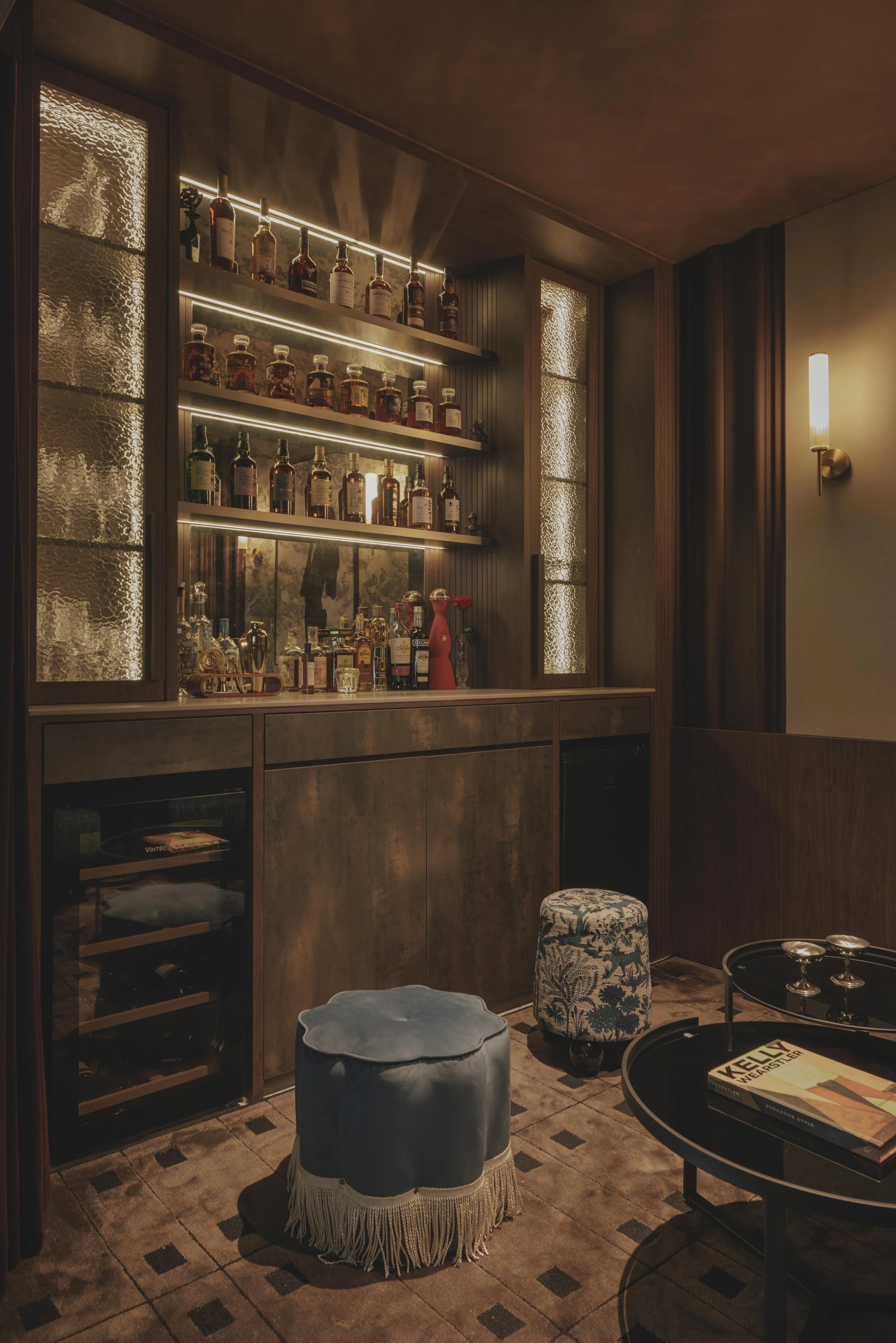 A luxurious home bar area with shelves of various liquor bottles illuminated by warm lighting, a textured glass wine fridge, and elegant furniture including a patterned stool and a round black table with magazines and glassware. The room has dark wood accents, textured walls, and a cozy ambiance.