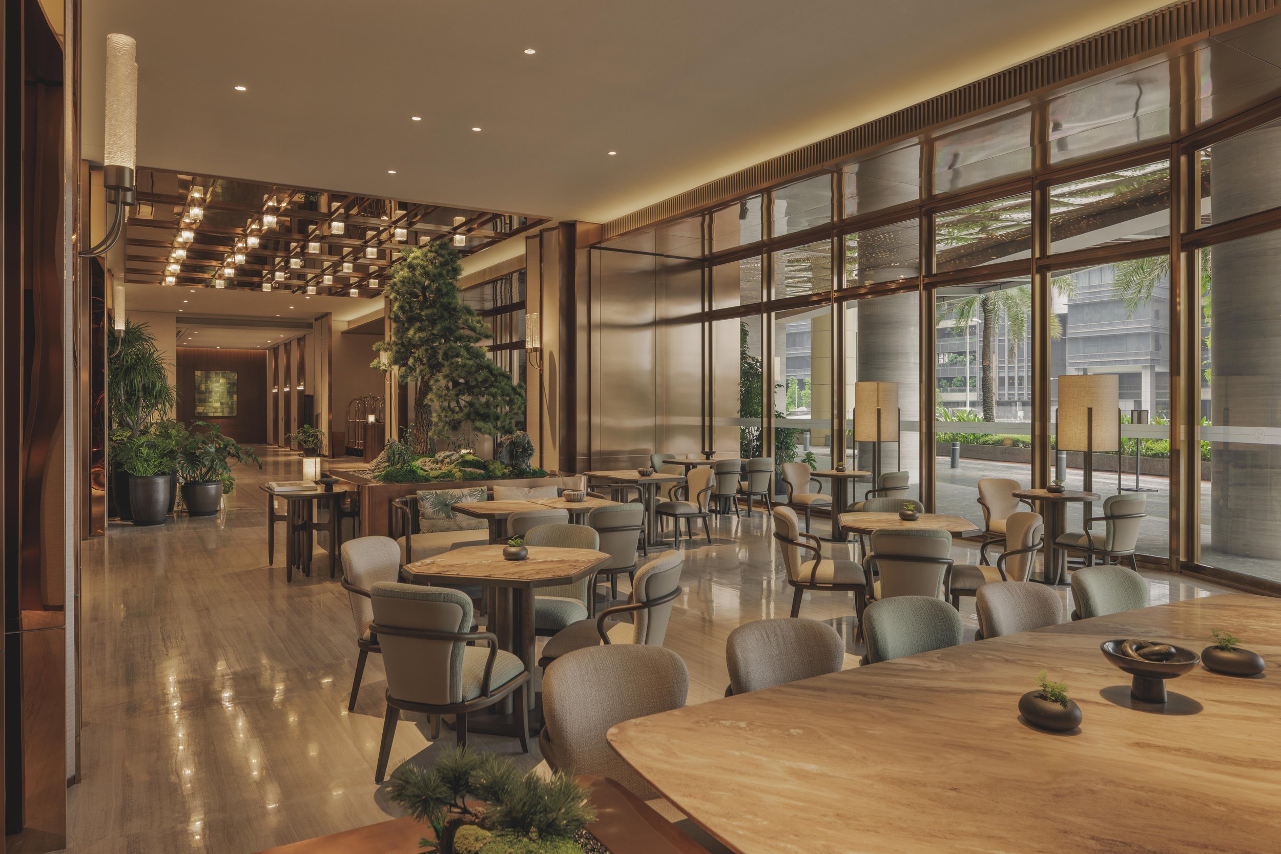 Interior view of a modern hotel lobby or lounge area with wooden tables, upholstered chairs, potted plants, large floor-to-ceiling windows, and decorative lighting fixtures.