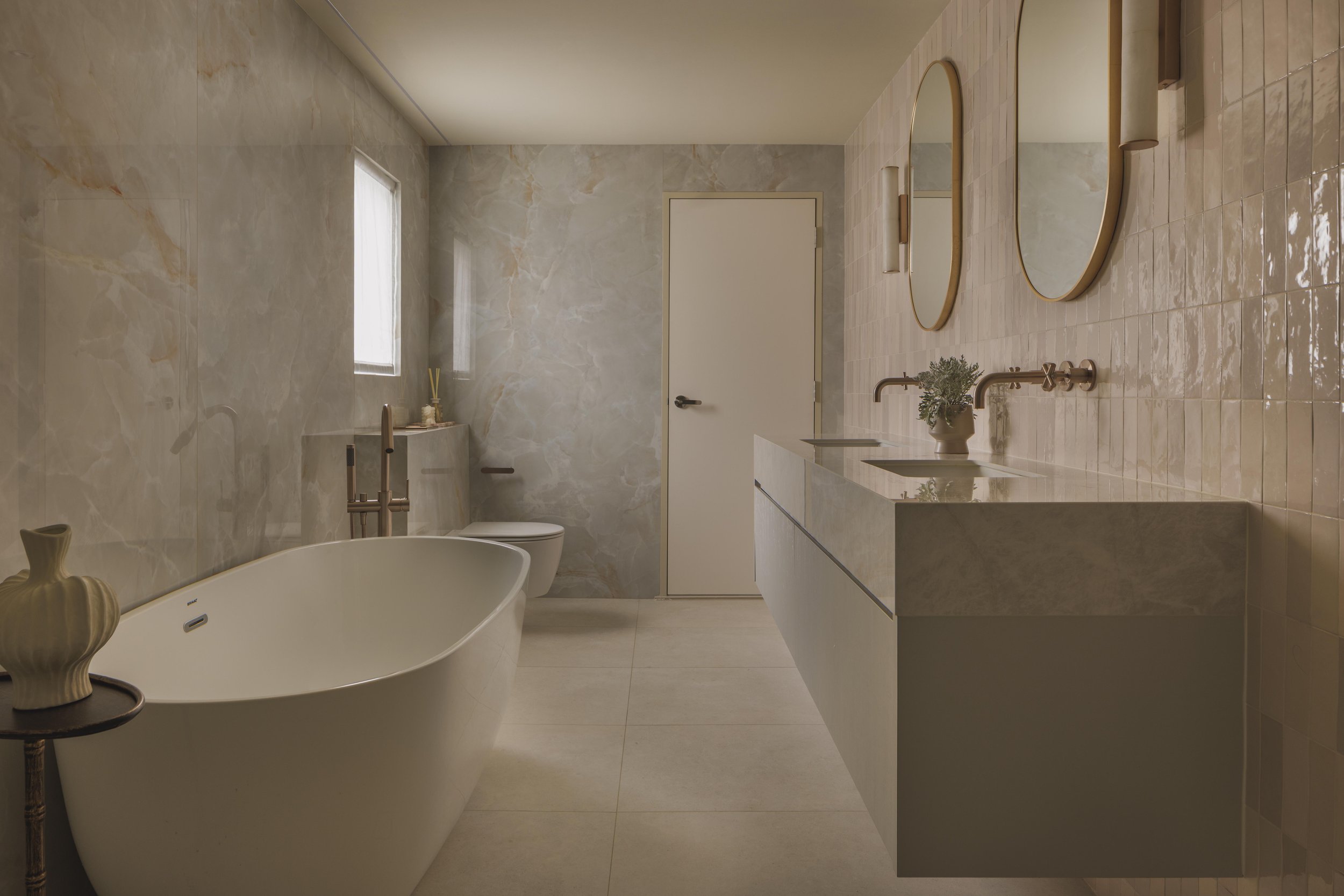 Modern bathroom with beige marble walls and floor, a freestanding bathtub, a white vanity with two round mirrors above, a potted plant, and a closed door near a small window.