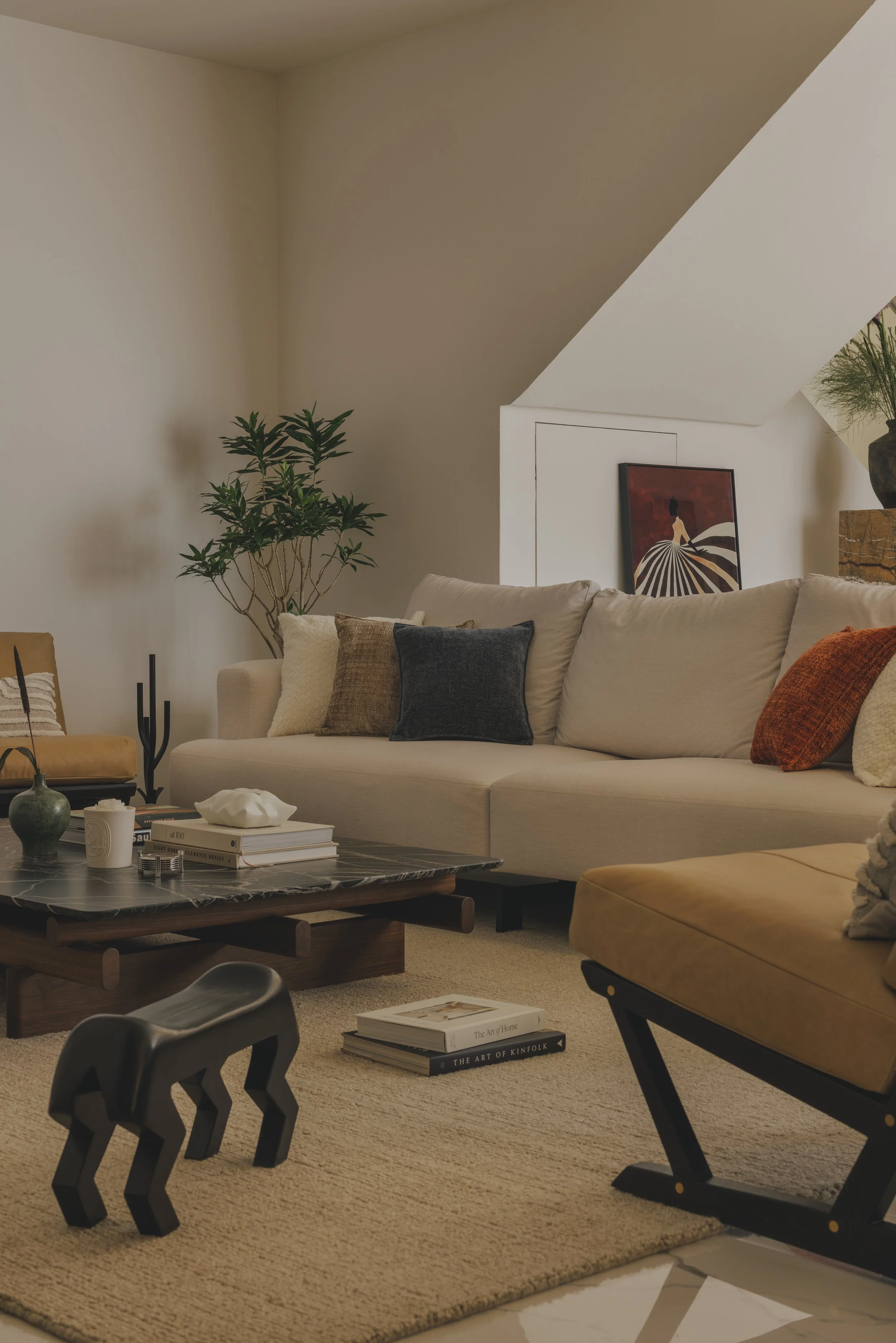 Living room with beige sofa adorned with throw pillows, a black marble coffee table with books and decorative objects, a yellow armchair, a large potted plant, wall art featuring a woman in a striped dress, and a beige carpet.