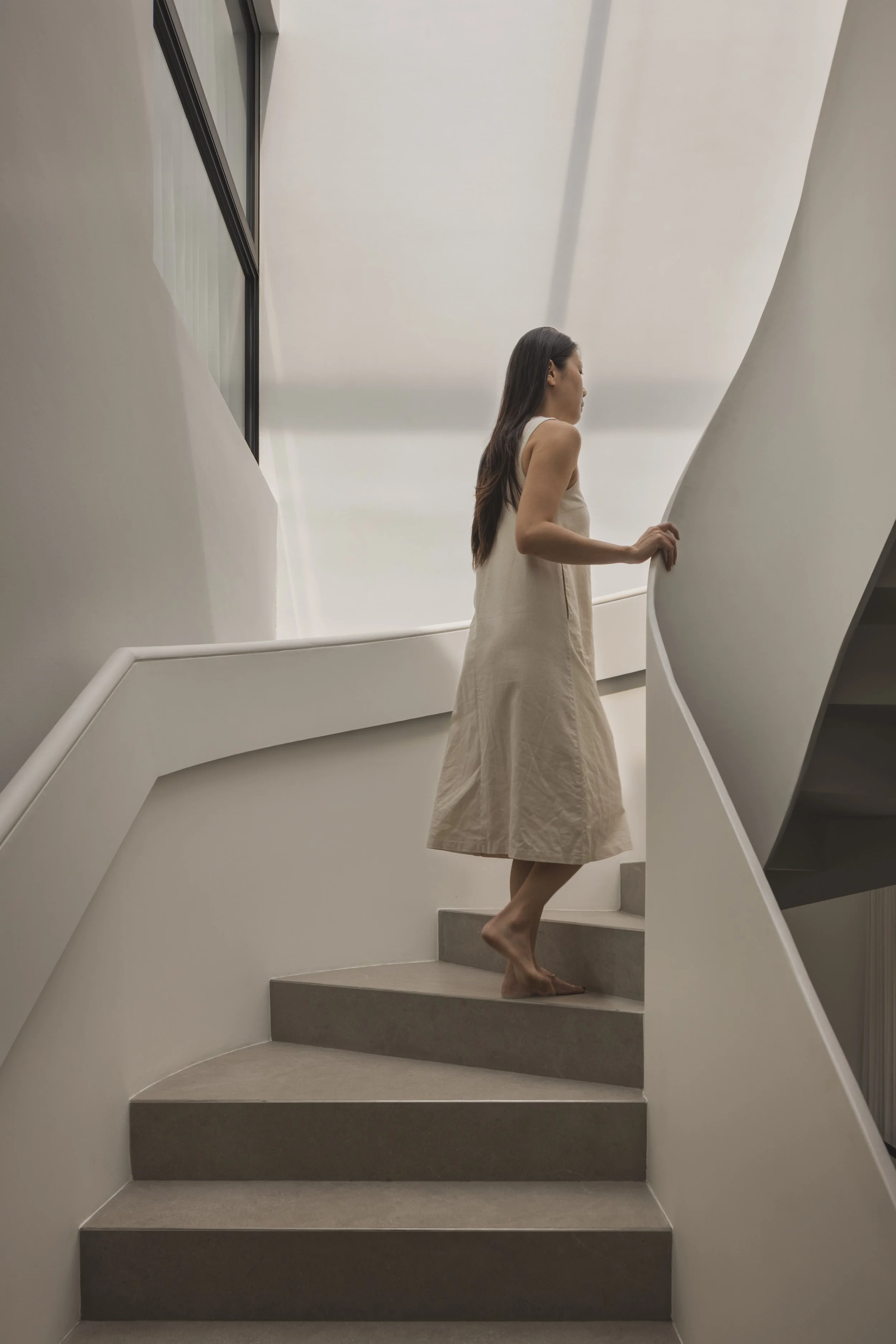 A woman with long dark hair walking barefoot up a curved staircase in a white modern building, wearing a beige dress.