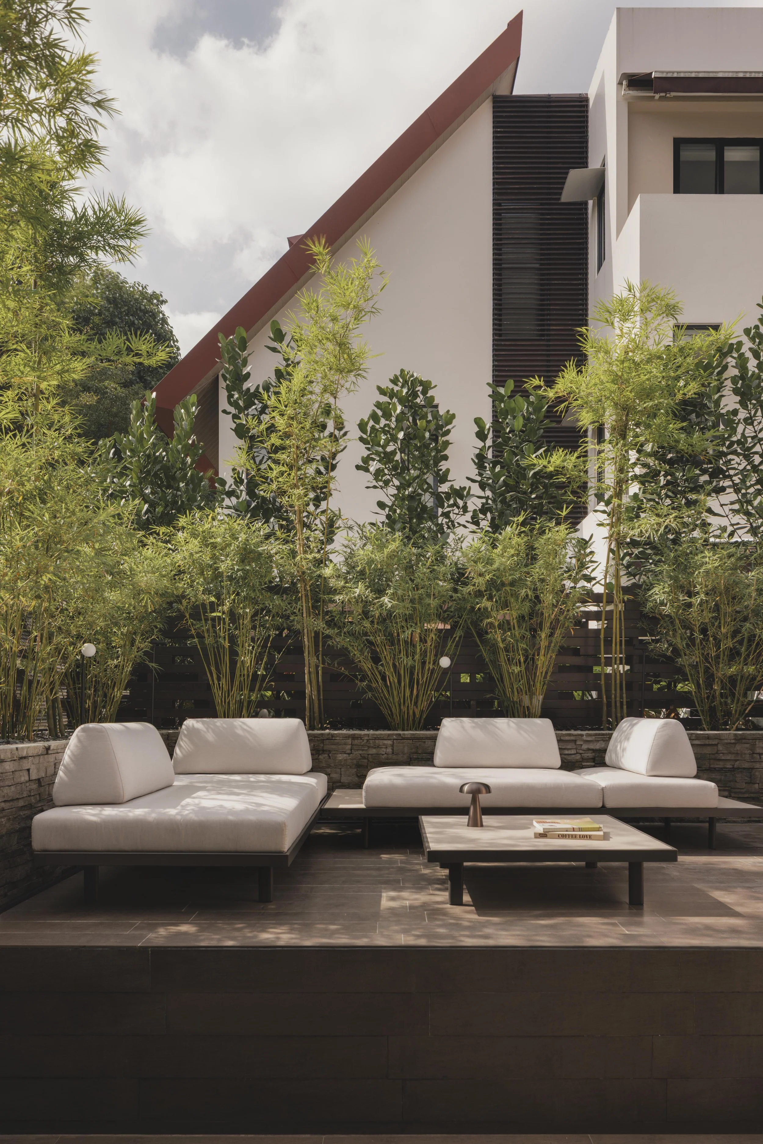 Outdoor patio with white modern sectional sofa, coffee table with books, lush green trees, and a building with black window frames and wooden slats.