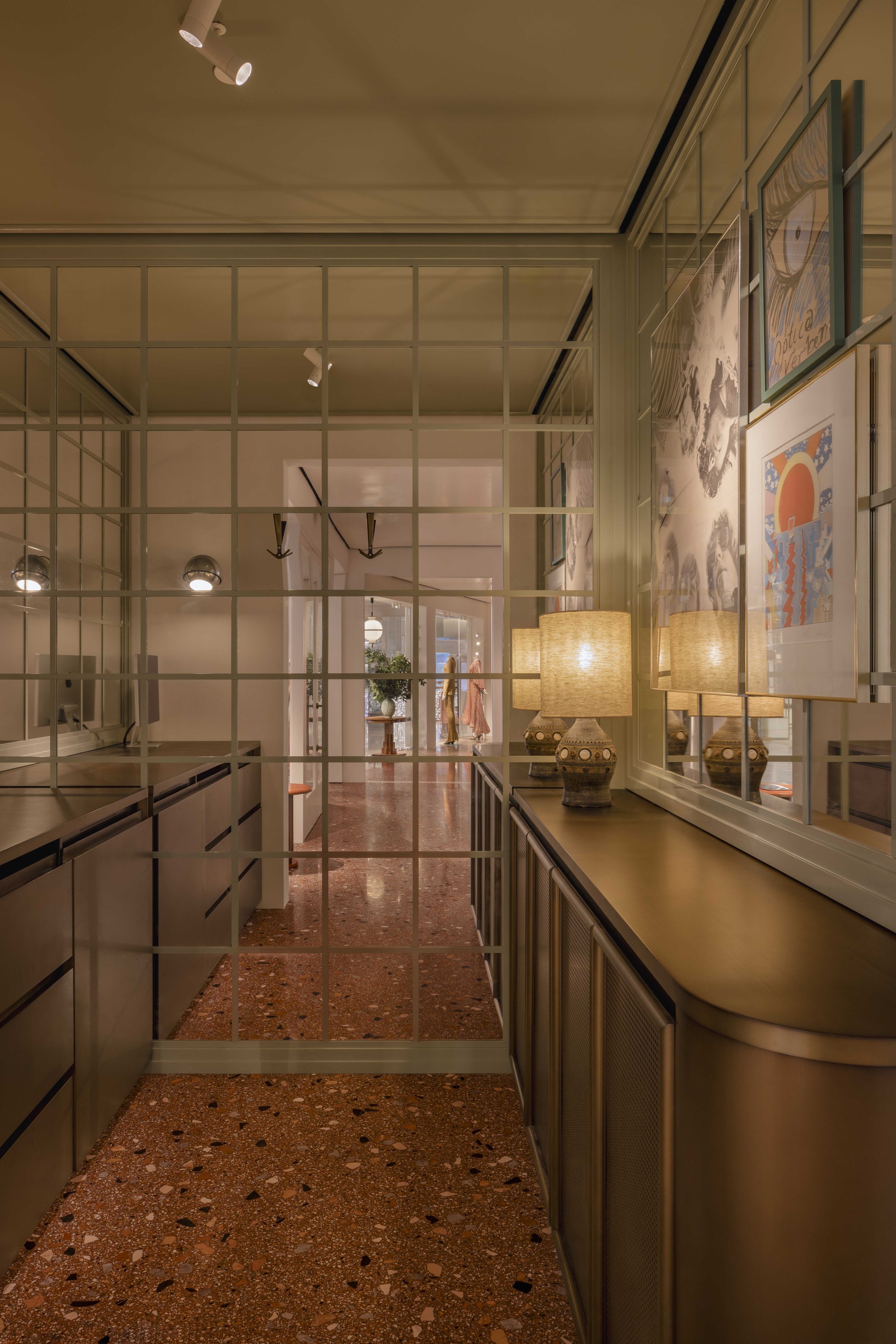 Interior view of a hallway with a glass partition, framed art on the wall, and lamps on a cabinet.