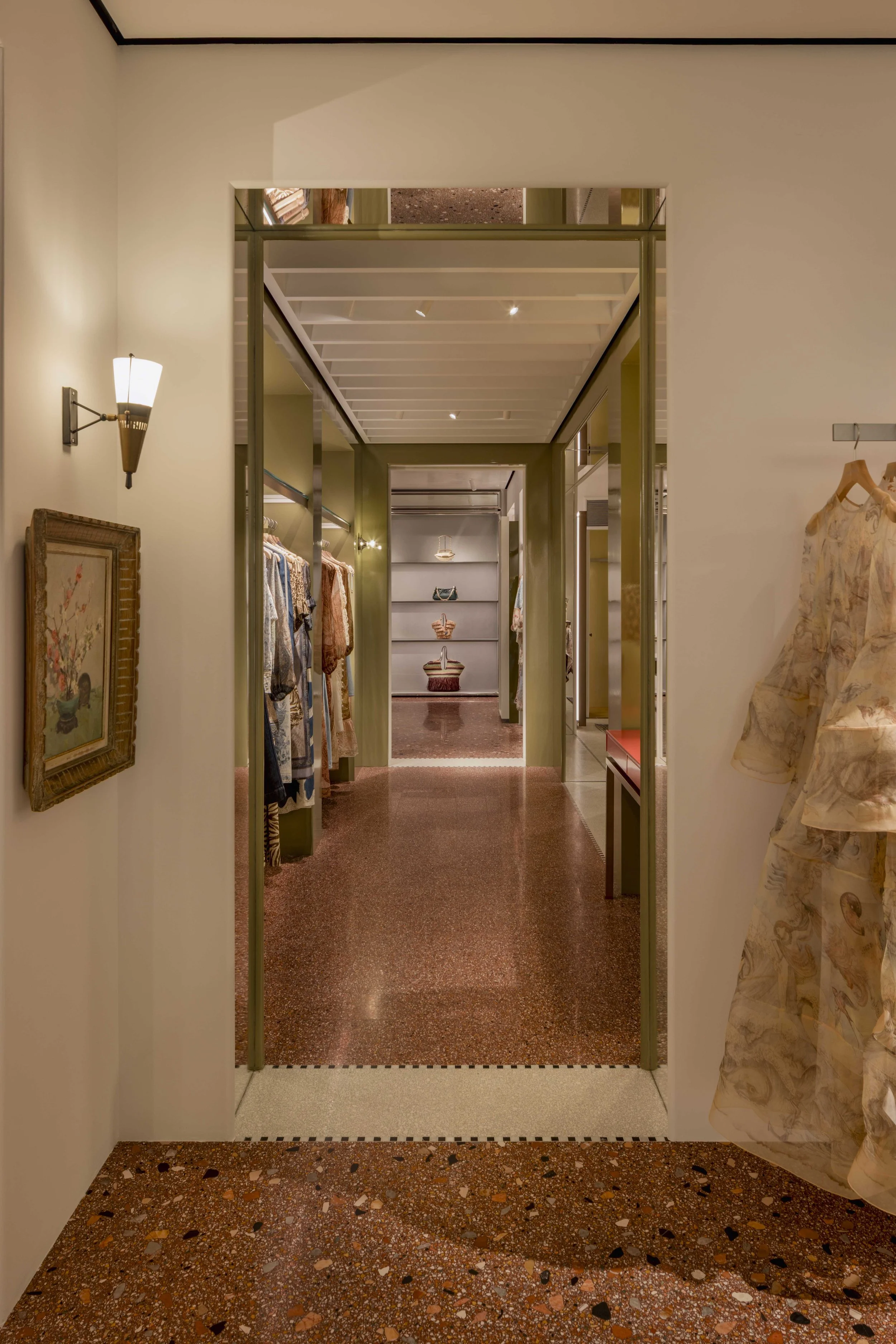 Interior view of a clothing boutique with a corridor leading to a back section, featuring clothing racks, framed artwork, and patterned flooring.