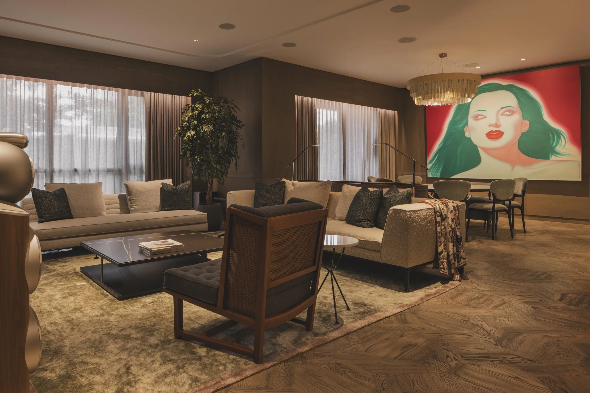 A luxurious hotel lounge with beige sofas, armchairs, and a coffee table on a patterned rug. Tall windows with sheer curtains let in natural light. A large colorful portrait of a woman with green hair and red lips adorns the wall. A chandelier hangs from the ceiling, and a potted plant adds greenery to the space.