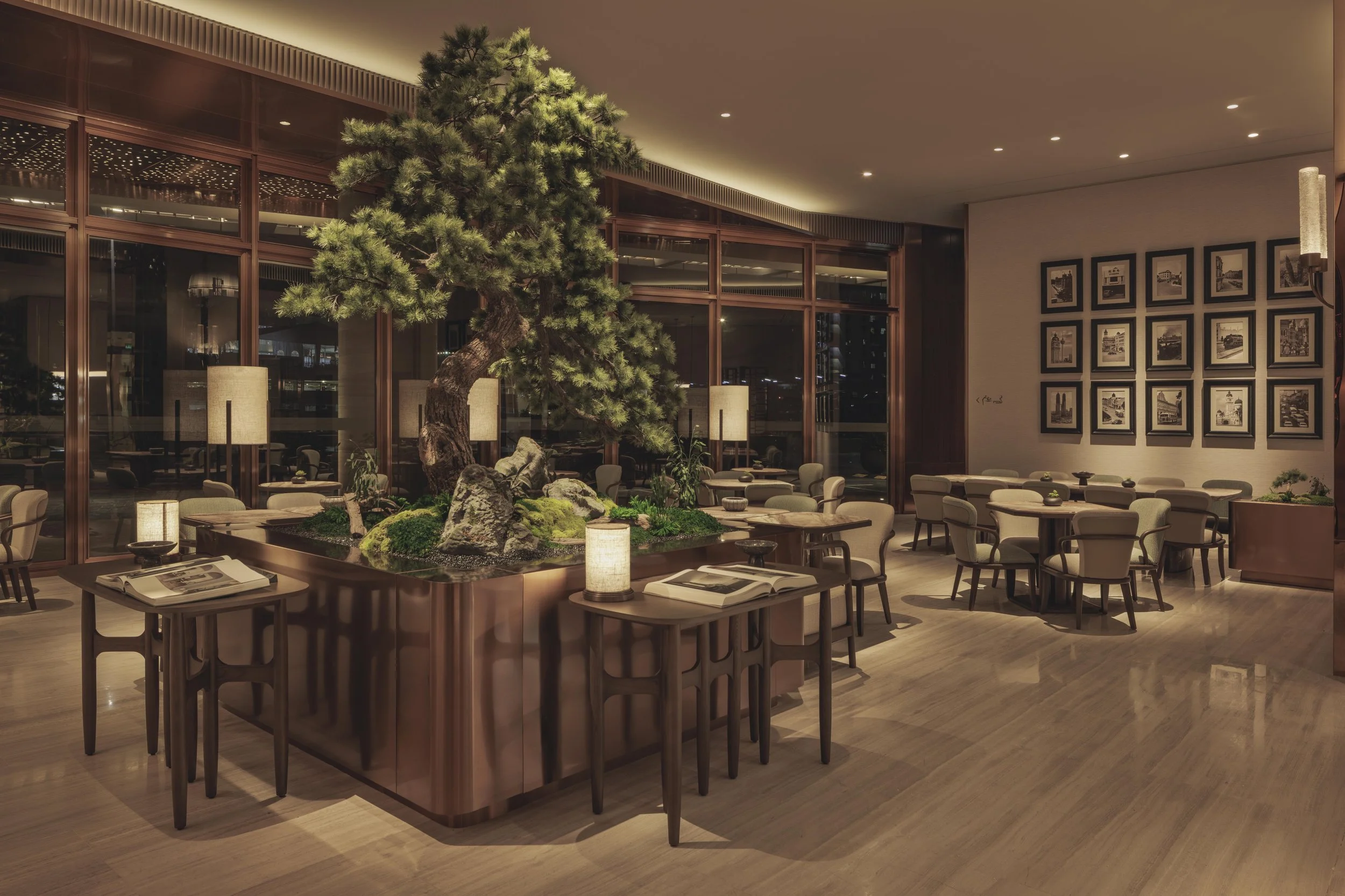 A modern indoor restaurant featuring a large decorative pine tree surrounded by rocks and greenery, with a city view outside through large glass windows. The space has a warm lighting and has framed black-and-white photographs on the walls, beige chairs around tables, and wood flooring.
