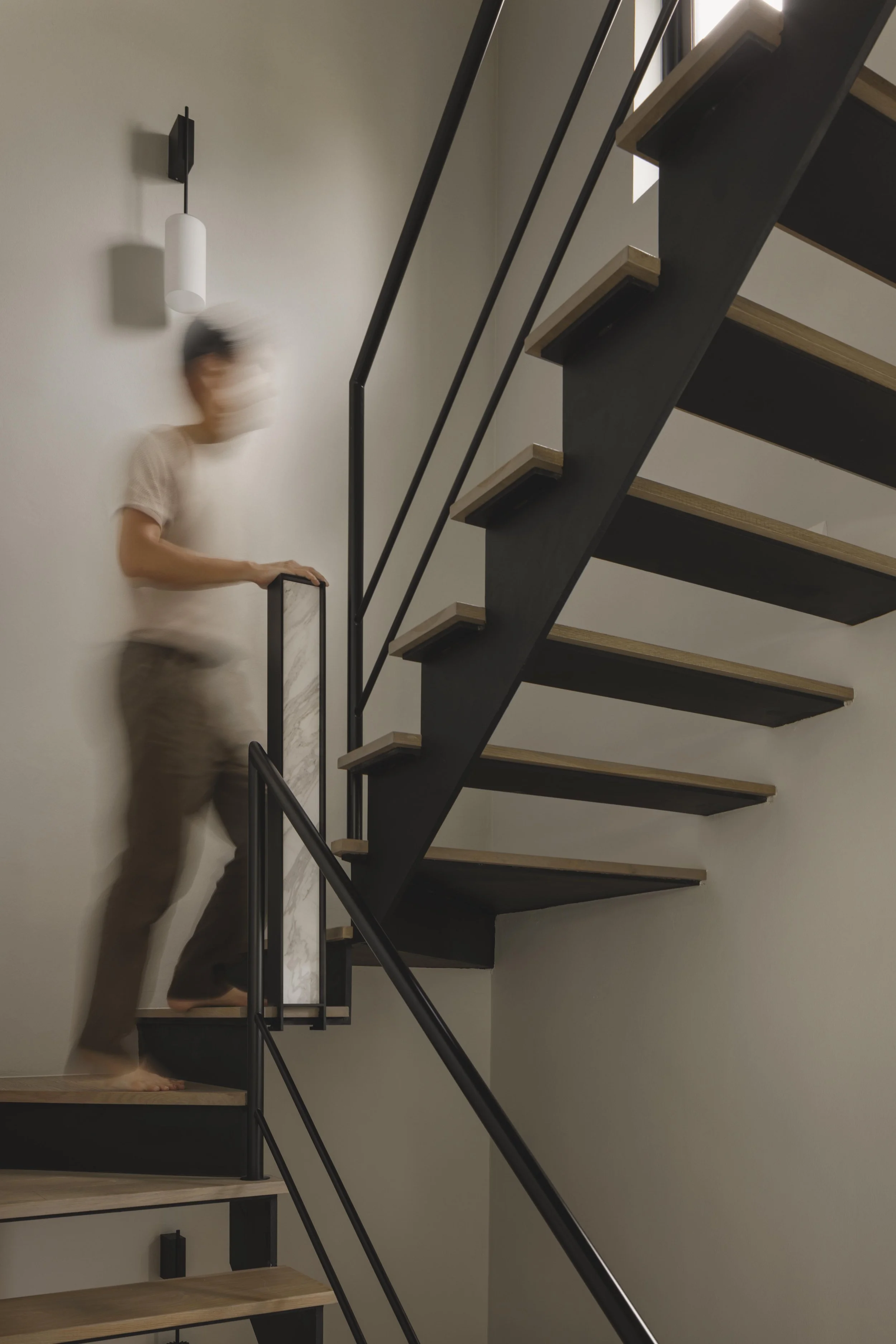 A blurred person, wearing a mask and a beige shirt, walking on a modern staircase with wooden steps and black metal railings, inside a minimalist interior space.