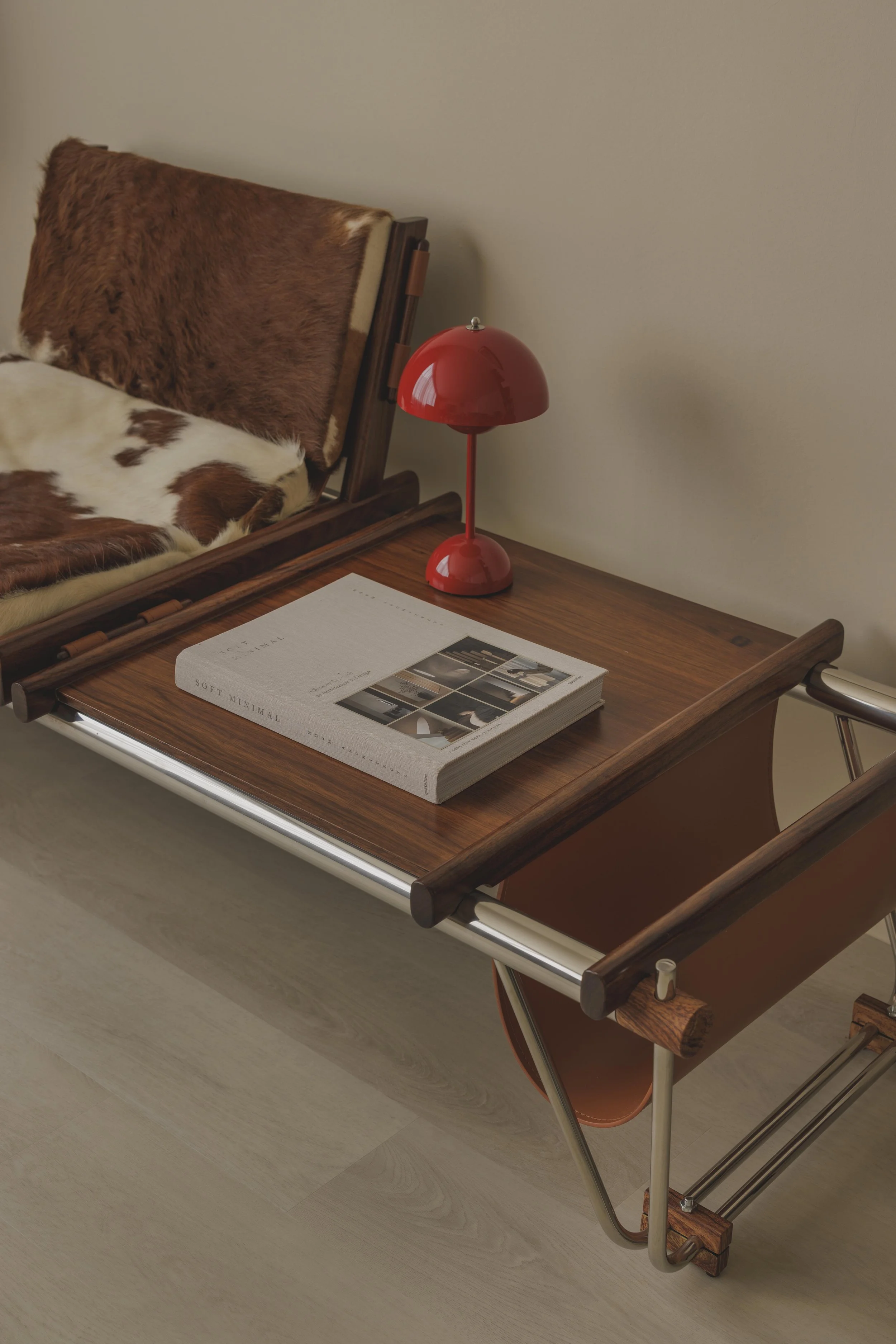A mid-century modern wooden bed with cowhide and faux fur pillows and throws, a red table lamp, and an open magazine on a wooden bedside table.