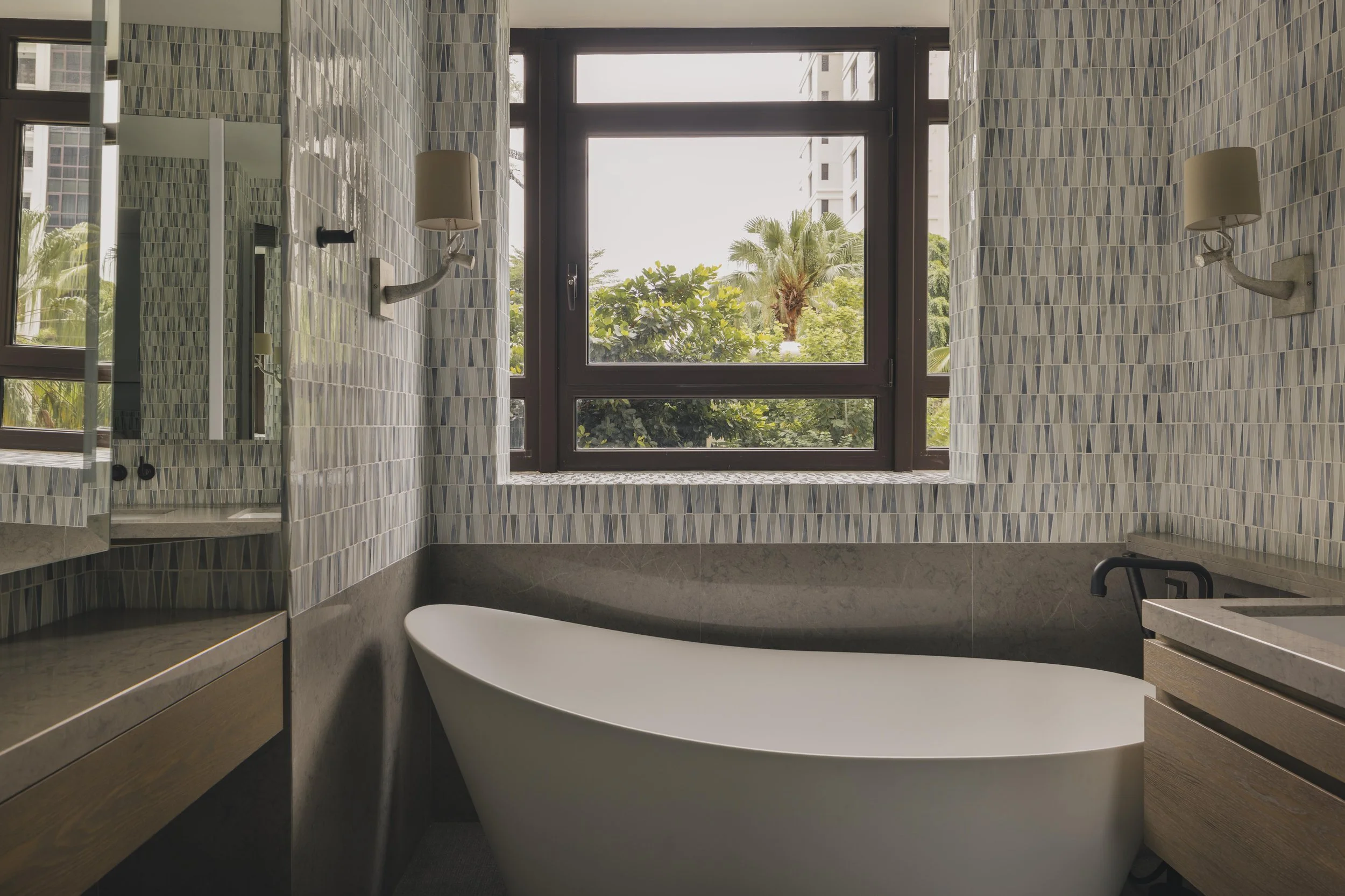 Modern bathroom with a freestanding bathtub beneath a large window, tiled walls with a geometric pattern, and tropical trees visible outside.