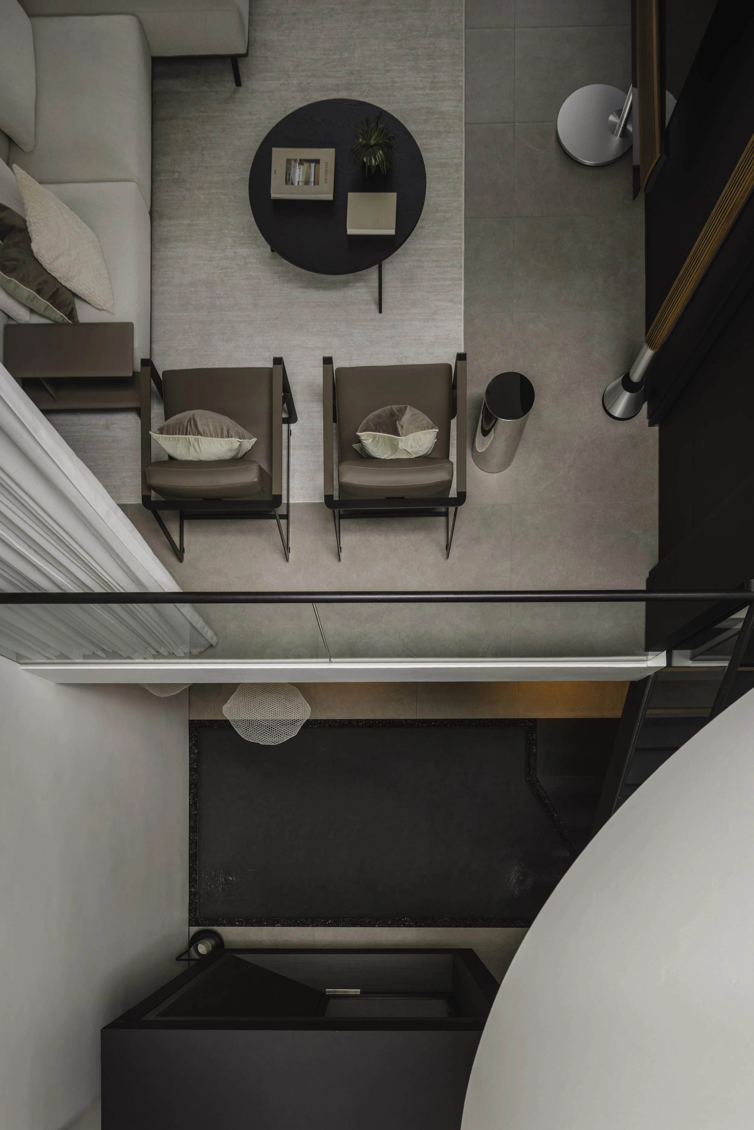 Top-down view of a living area with a beige sofa, two brown armchairs with pillows, a round black coffee table with books and a plant, and a tiled floor transition to a darker entryway with a black door and a gray mat.