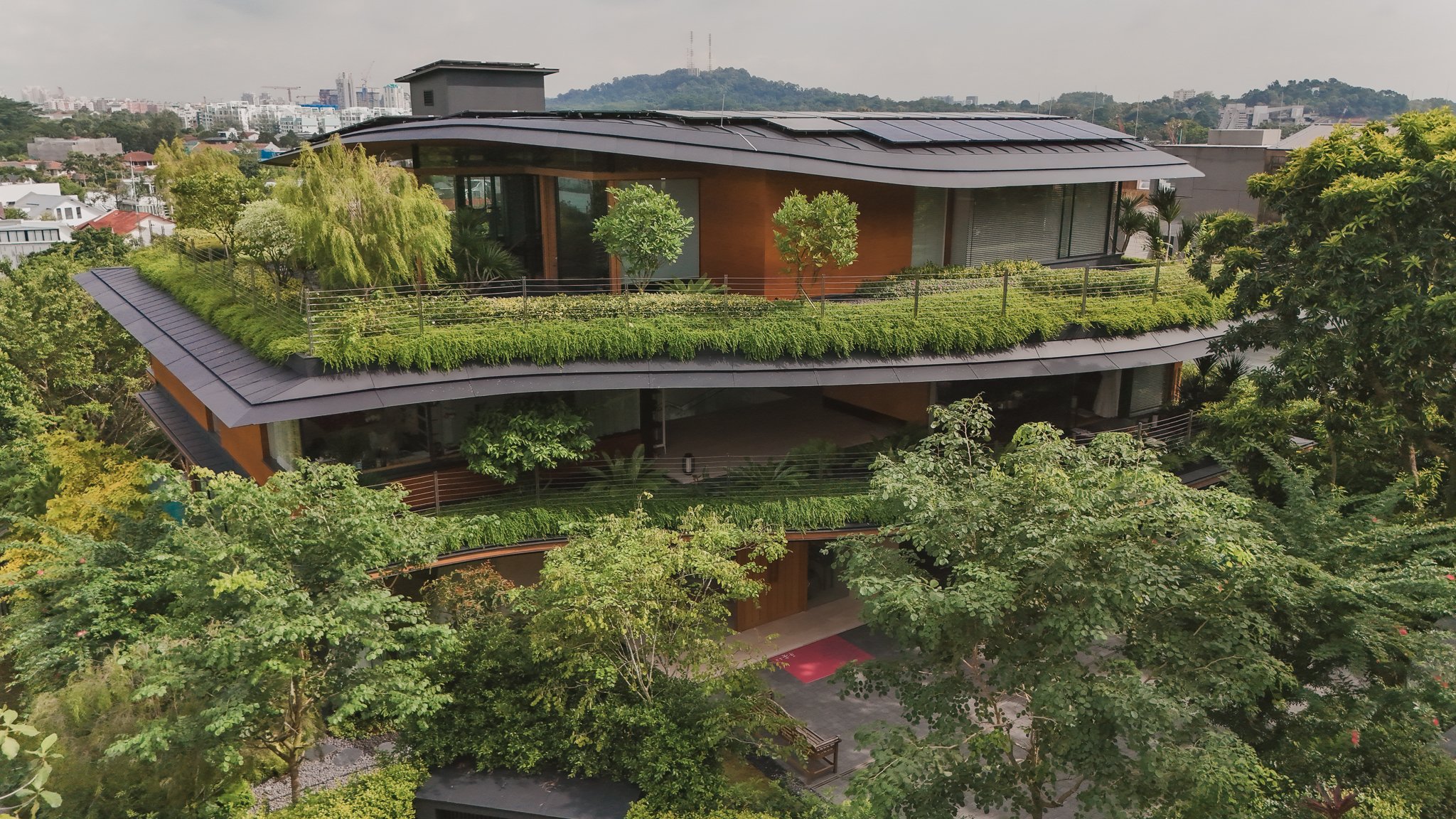 A modern multi-story house with a green rooftop garden, surrounded by trees and overlooking a cityscape in the background.