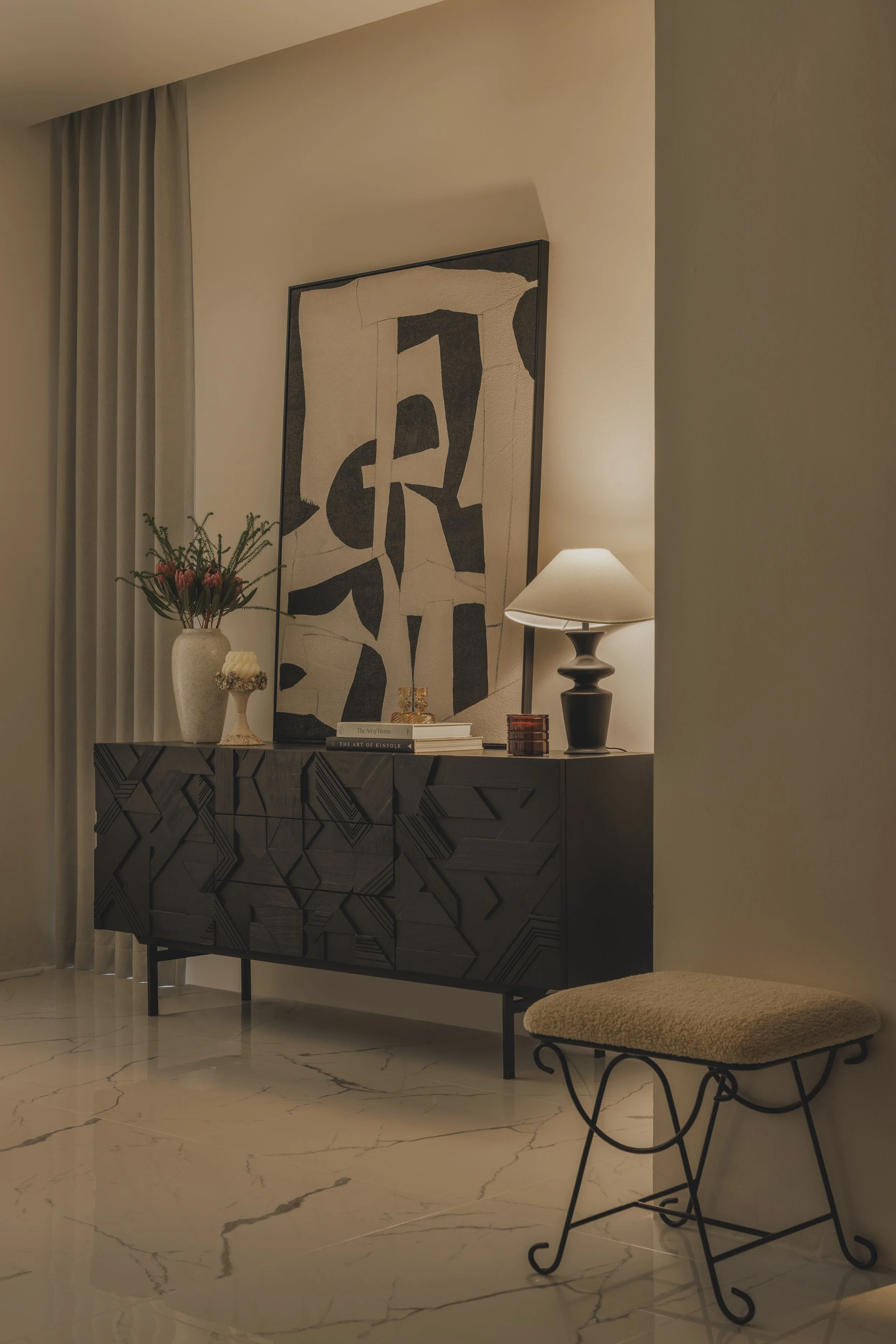 A corner of a living room with a decorative black and white cabinet, a tall vase with pink flowers, a small white candle holder, and three books. Above the cabinet hangs a large abstract black and white artwork. To the right, there is a black table lamp with a beige shade and a beige upholstered bench with curled black metal legs. The room has light-colored walls, marble flooring, and curtains on the left.