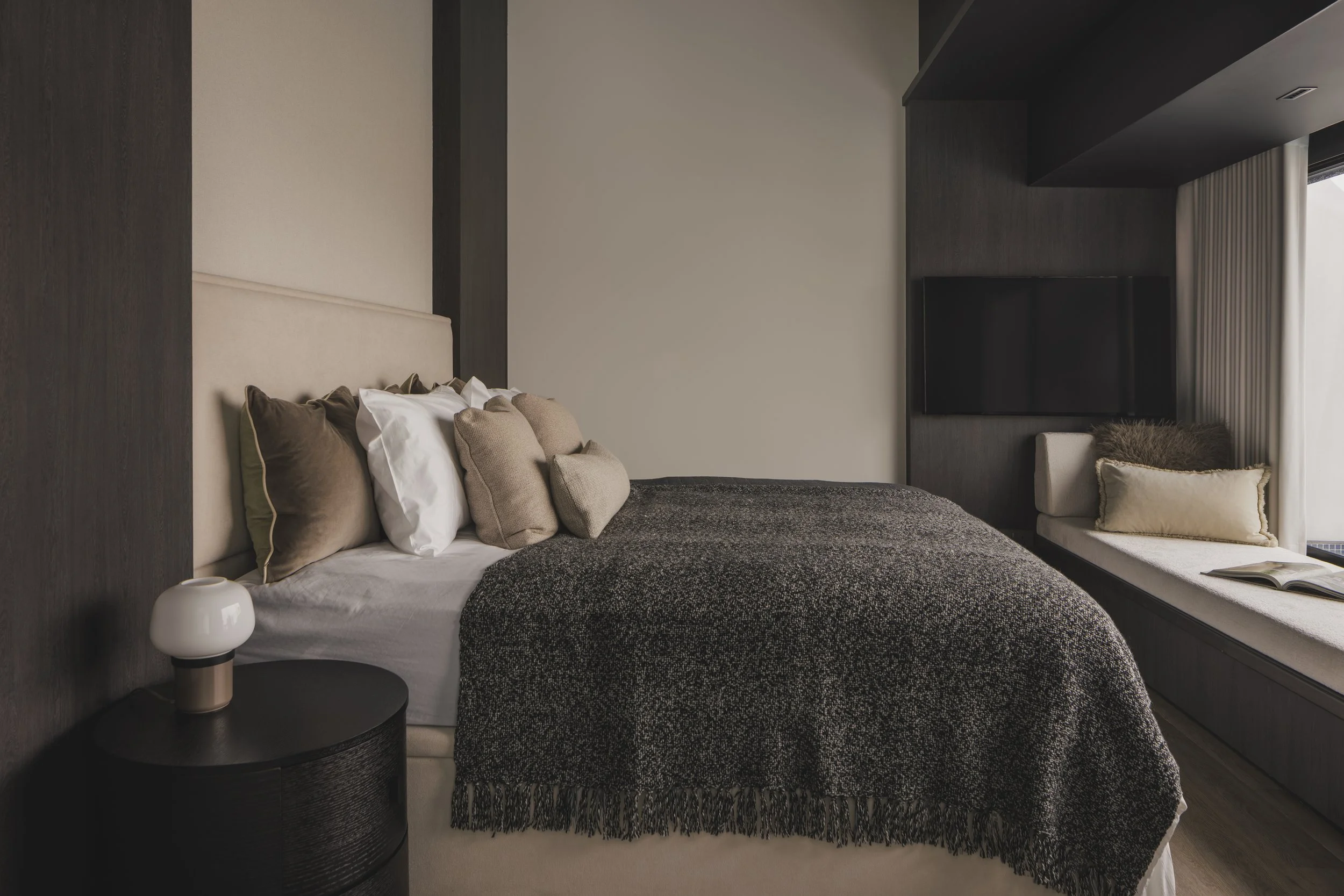Modern bedroom with a bed that has pillows, a dark textured blanket, and a beige headboard. There is a white lamp on a black nightstand next to the bed and a cushioned window seat with an open book and a beige pillow. The wall is painted light, with dark wood panels and a flat-screen TV on the wall.