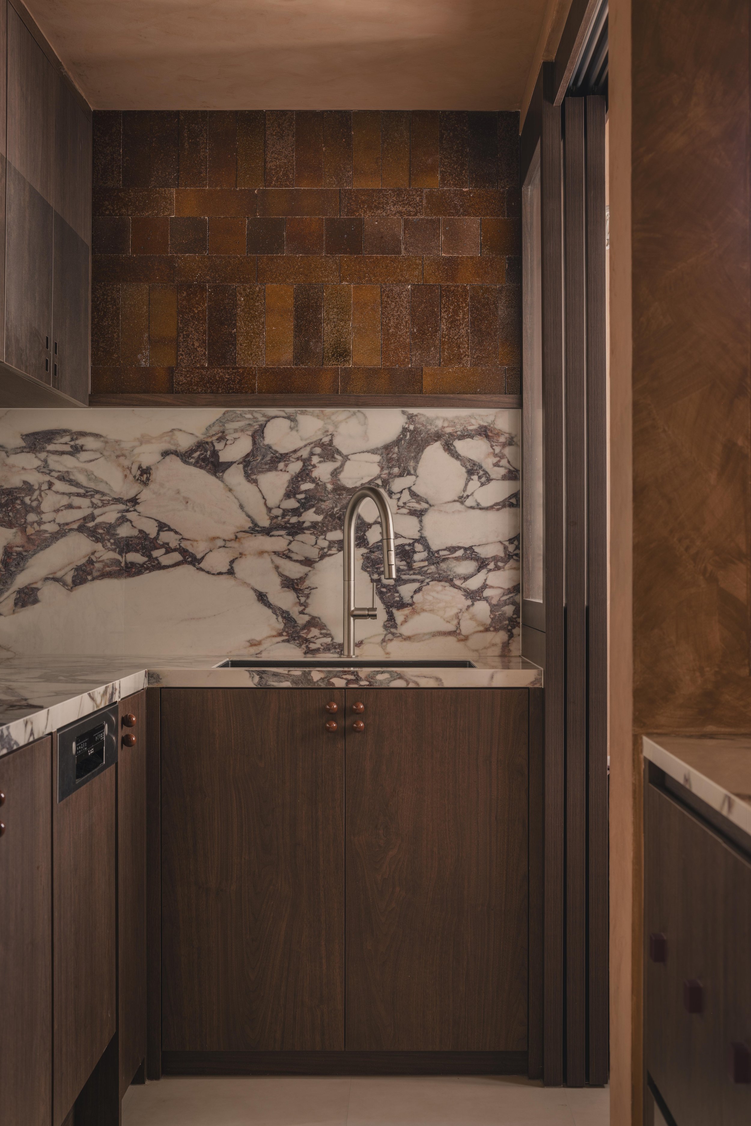 Kitchen with dark wood cabinetry, marble backsplash with black and brown veining, stainless steel faucet, and brown tiled flooring.