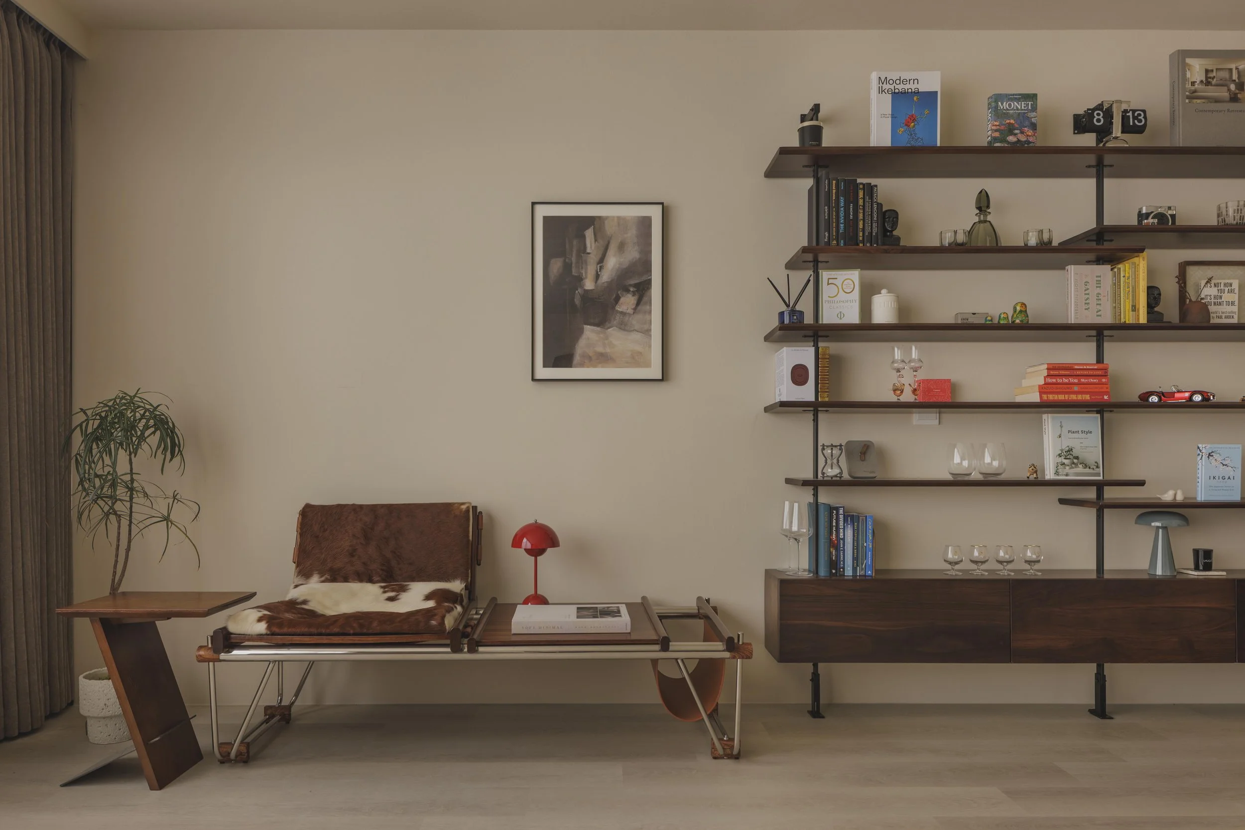 Interior of a living room with a beige wall, a wooden and metal bookshelf with decorative items, books, and framed art, a small table with a tray and a book, a LoveSac chair with a cowhide pillow, a red lamp, a potted plant, and a closed curtain on the left side.