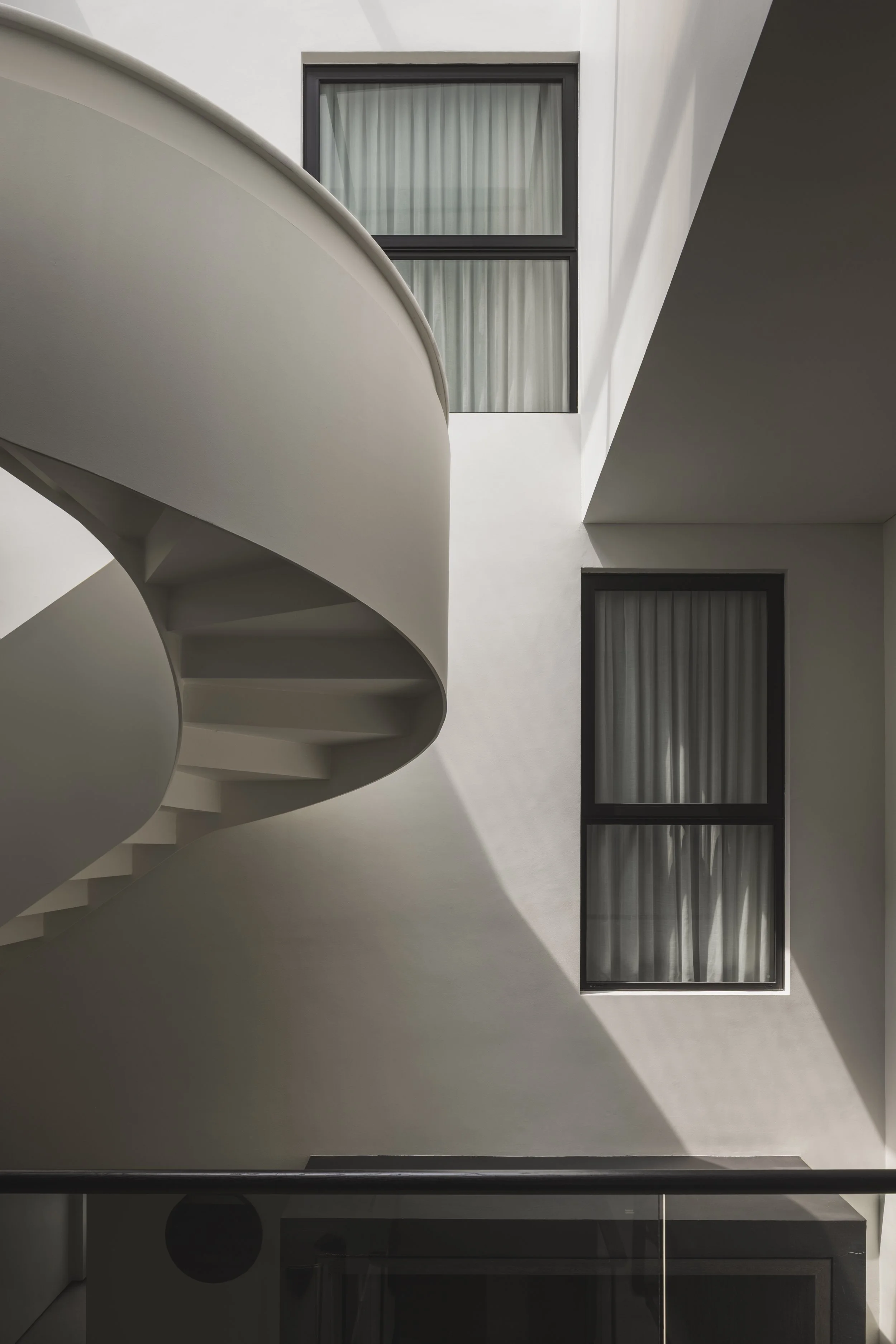 Minimalist modern building with white walls, black-framed windows with curtains, and a curved white staircase with shadows.