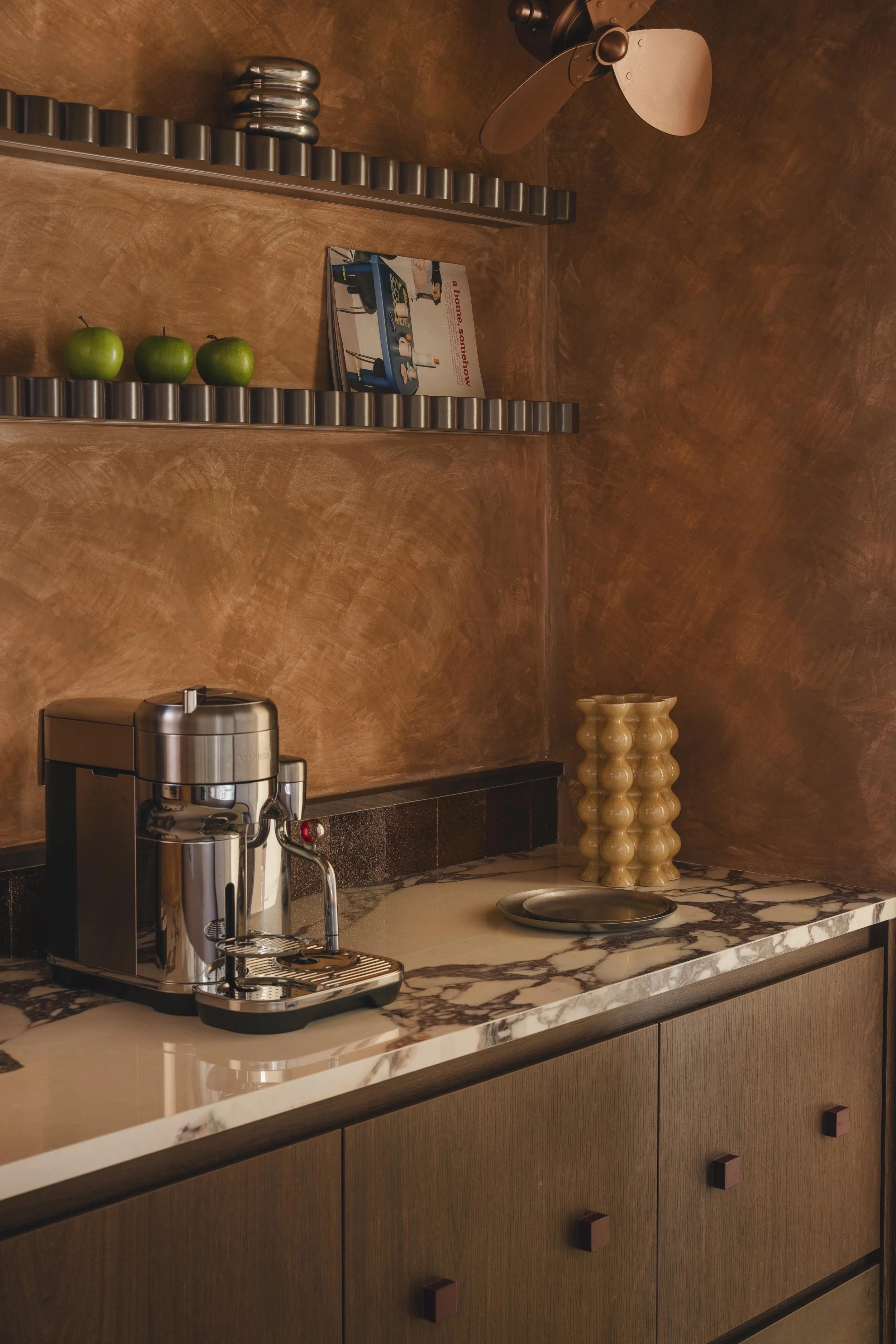 A kitchen countertop with a coffee machine, three green apples on a shelf, a magazine, and a beige decorative sculpture, with brown wooden cabinets and a marble surface.