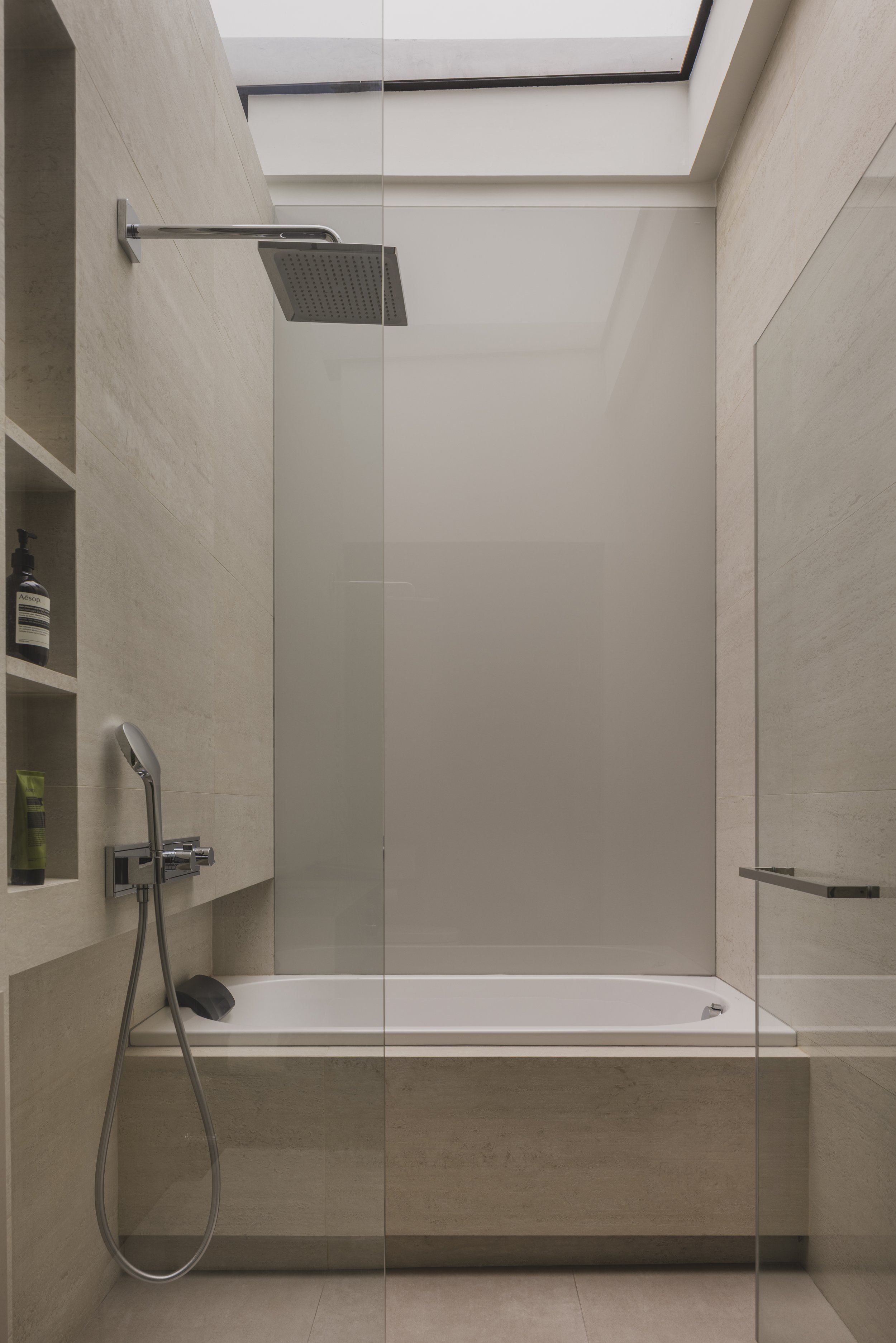 Modern walk-in shower with a rainfall showerhead, a handheld shower, and built-in shelves with toiletries, in a minimalist bathroom with beige tiles and a glass door.