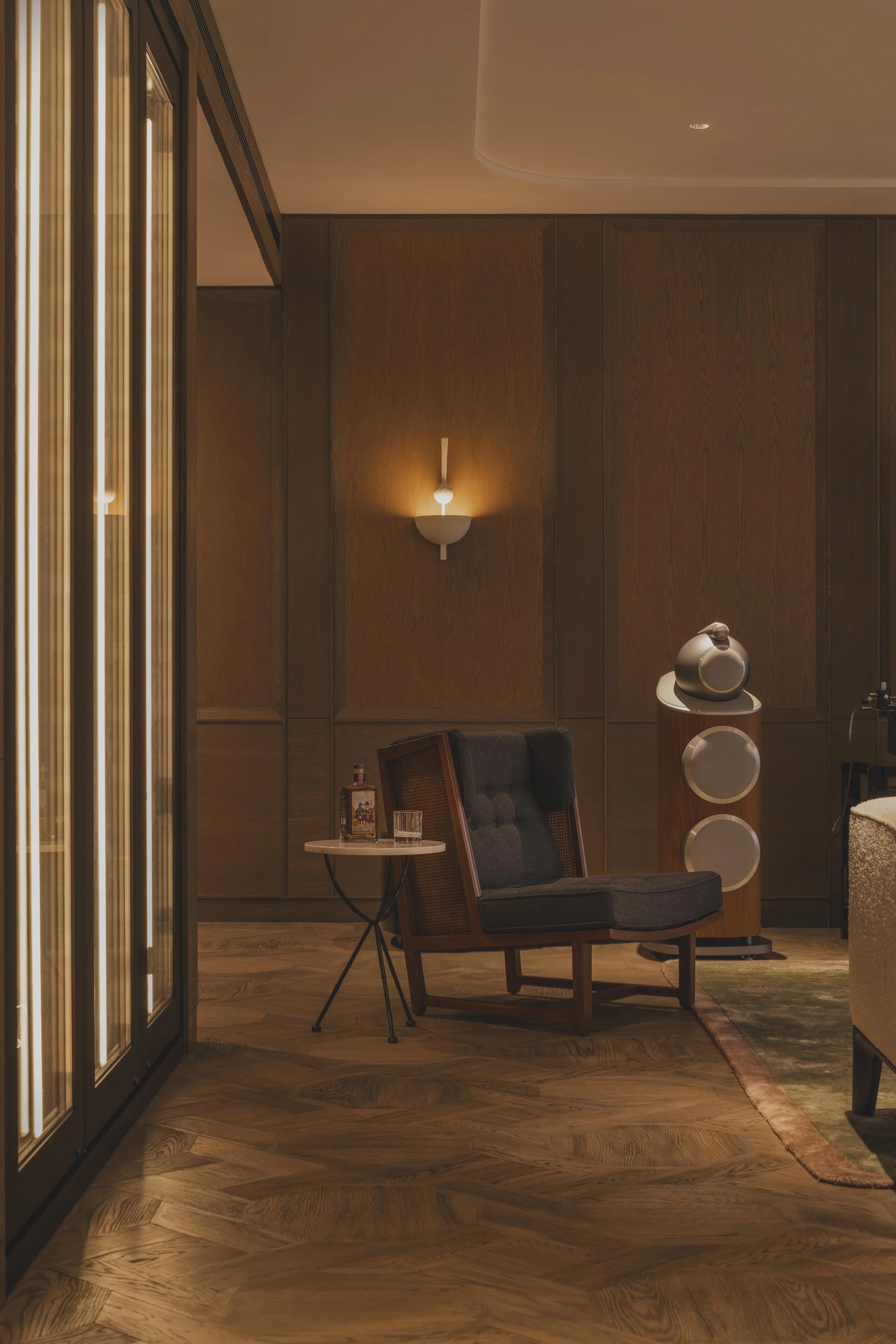 A cozy, warmly lit room with wooden walls and flooring, featuring a black armchair with a wooden frame, a small side table with drinks, a modern speaker, and a decorative rug. There is a wall-mounted light fixture and marble glasses on the table.