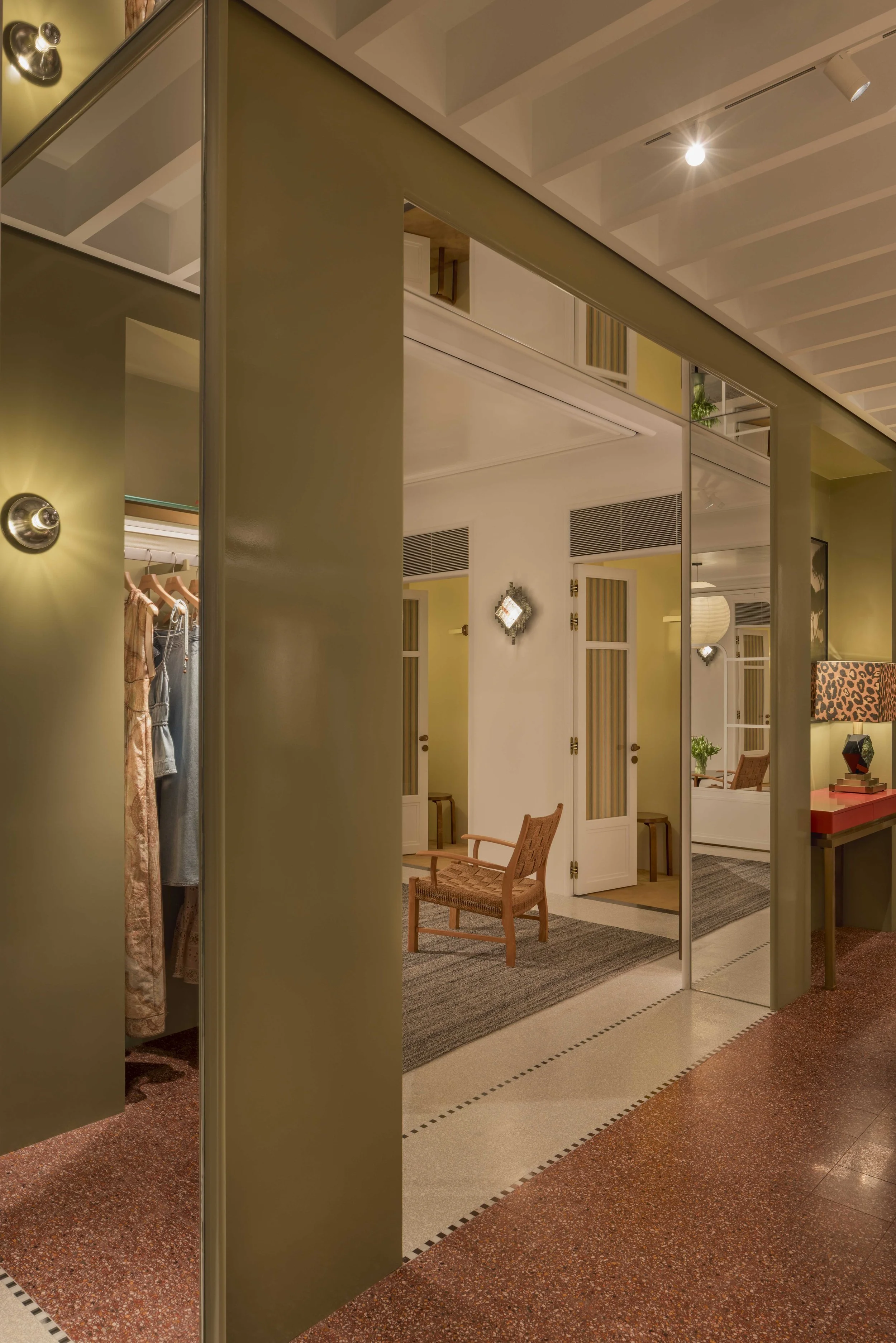 A modern indoor space with a closet, chairs, and a mirror, lit by ceiling spotlights and wall lamps, with a mix of beige, brown, and green tones.