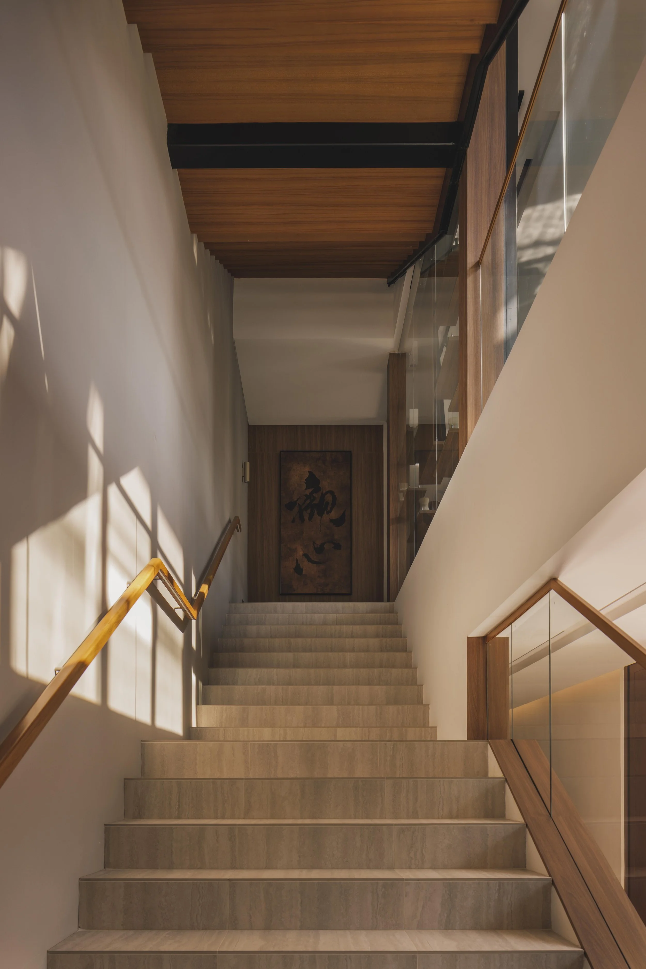 Interior view of a modern staircase with light-colored steps, wooden handrails, and glass panels on a staircase landing. Shadows of window frames are cast on the left wall, and there is a framed artwork or calligraphy on the back wall.