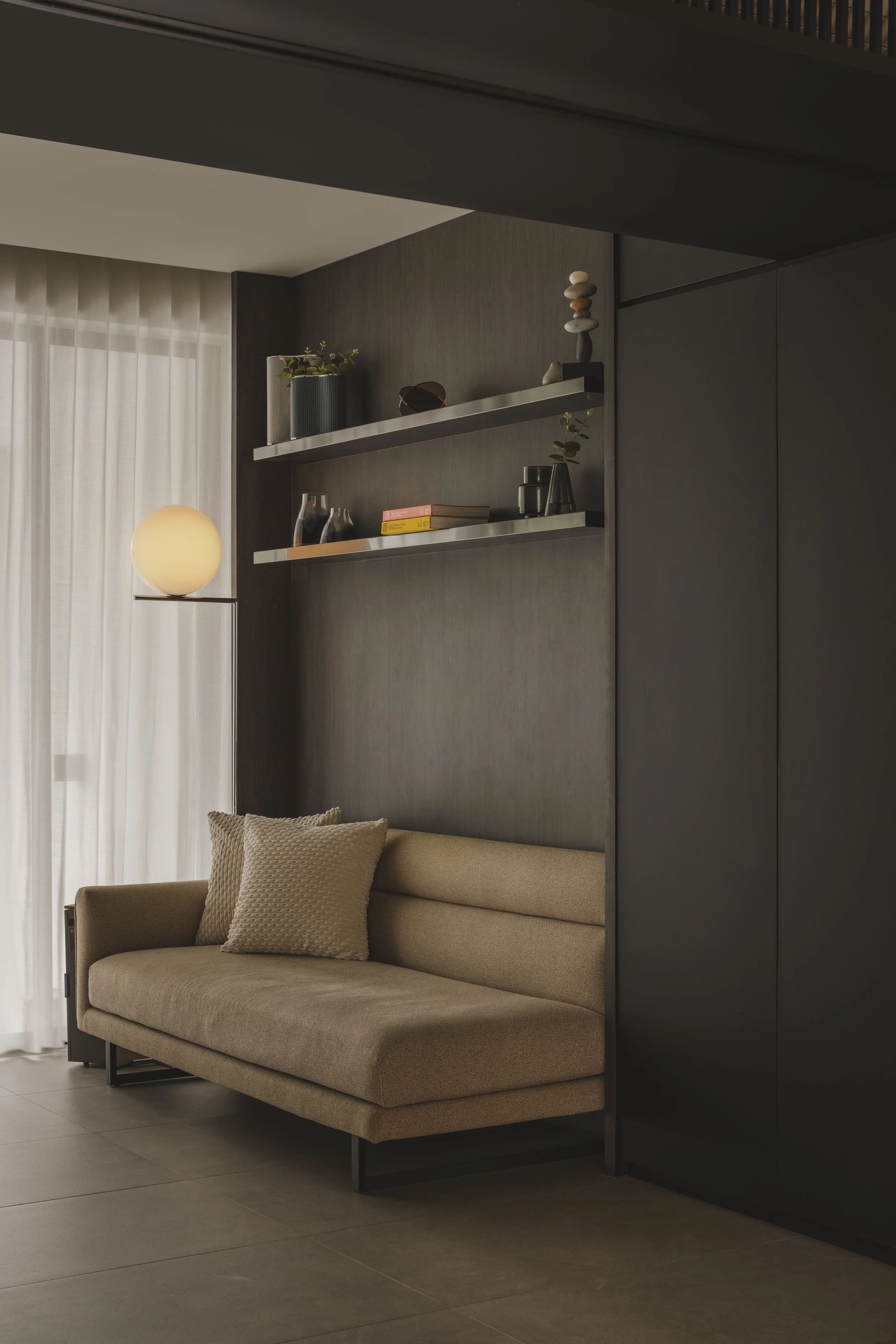 Modern living room with beige sofa and pillows, dark accent wall with floating shelves decorated with books, plants, and decorative objects, a spherical table lamp, and a large window with light curtains.