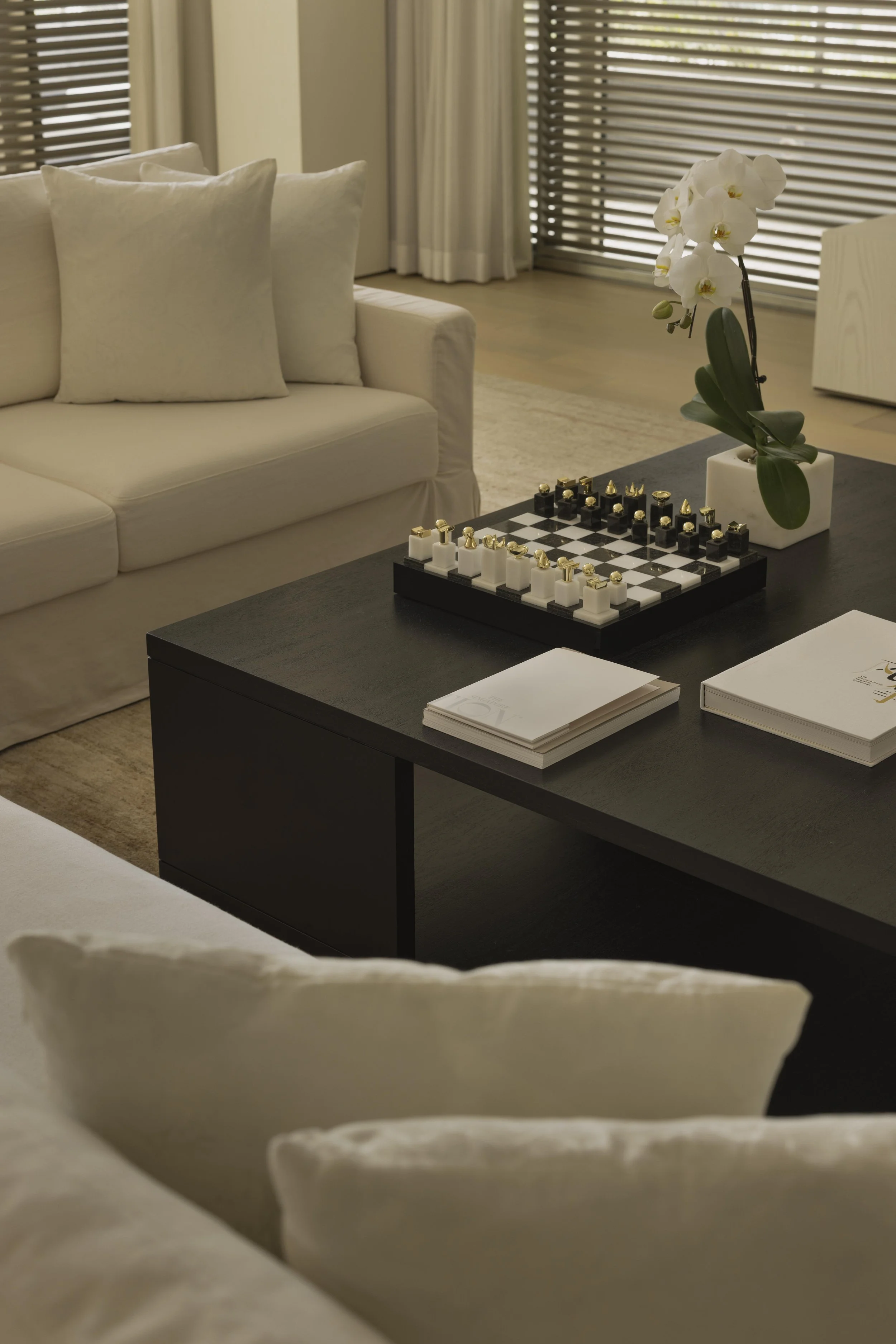 Living room with white sofa and cushions, a coffee table with a chess set, and a potted orchid plant, with wooden blinds on the windows and a light-colored rug.