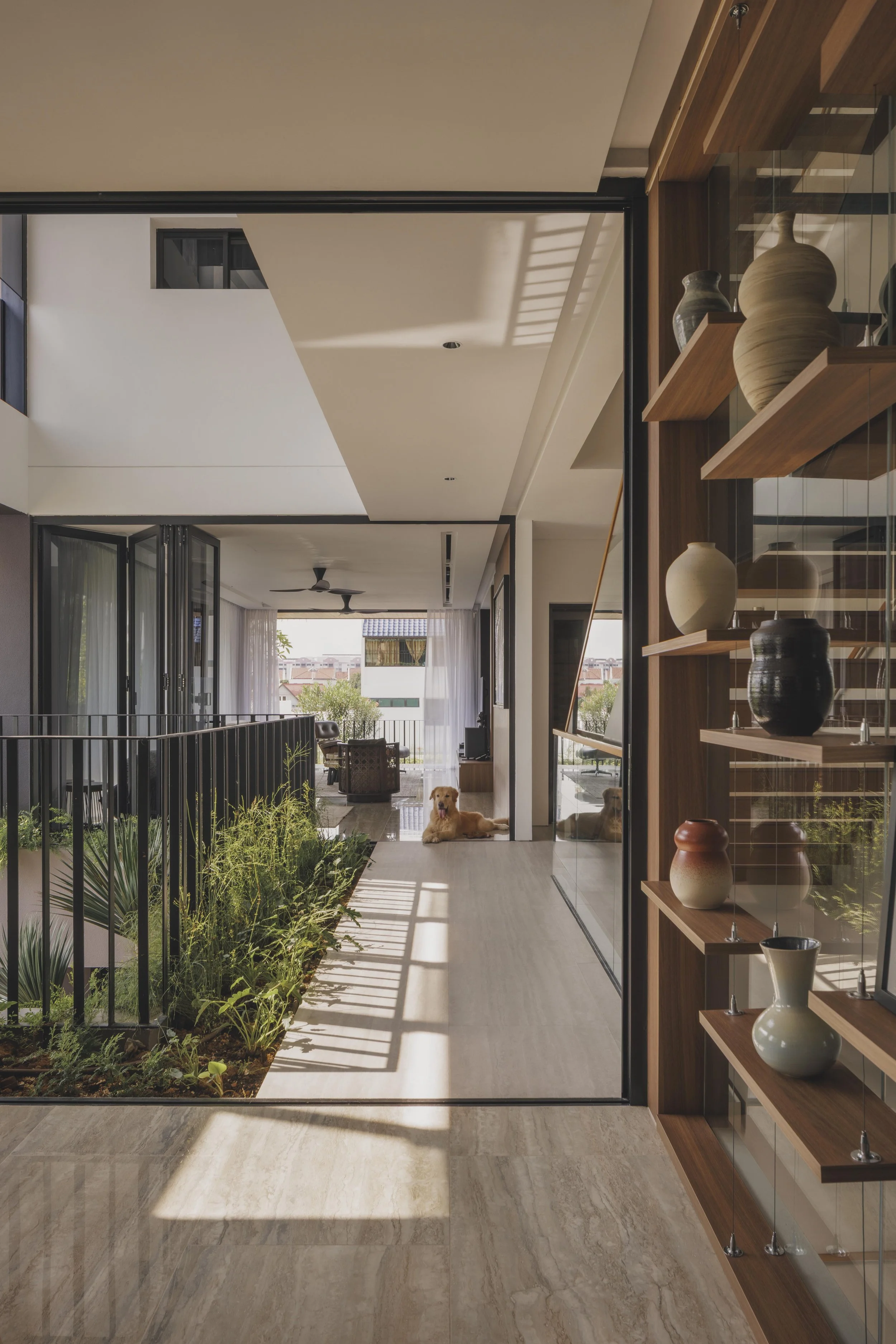 Modern interior hallway with large glass doors leading to an outdoor area, a wooden shelf with decorative vases on the right side, and a dog resting on the floor near the windows, with plants and outdoor view visible.