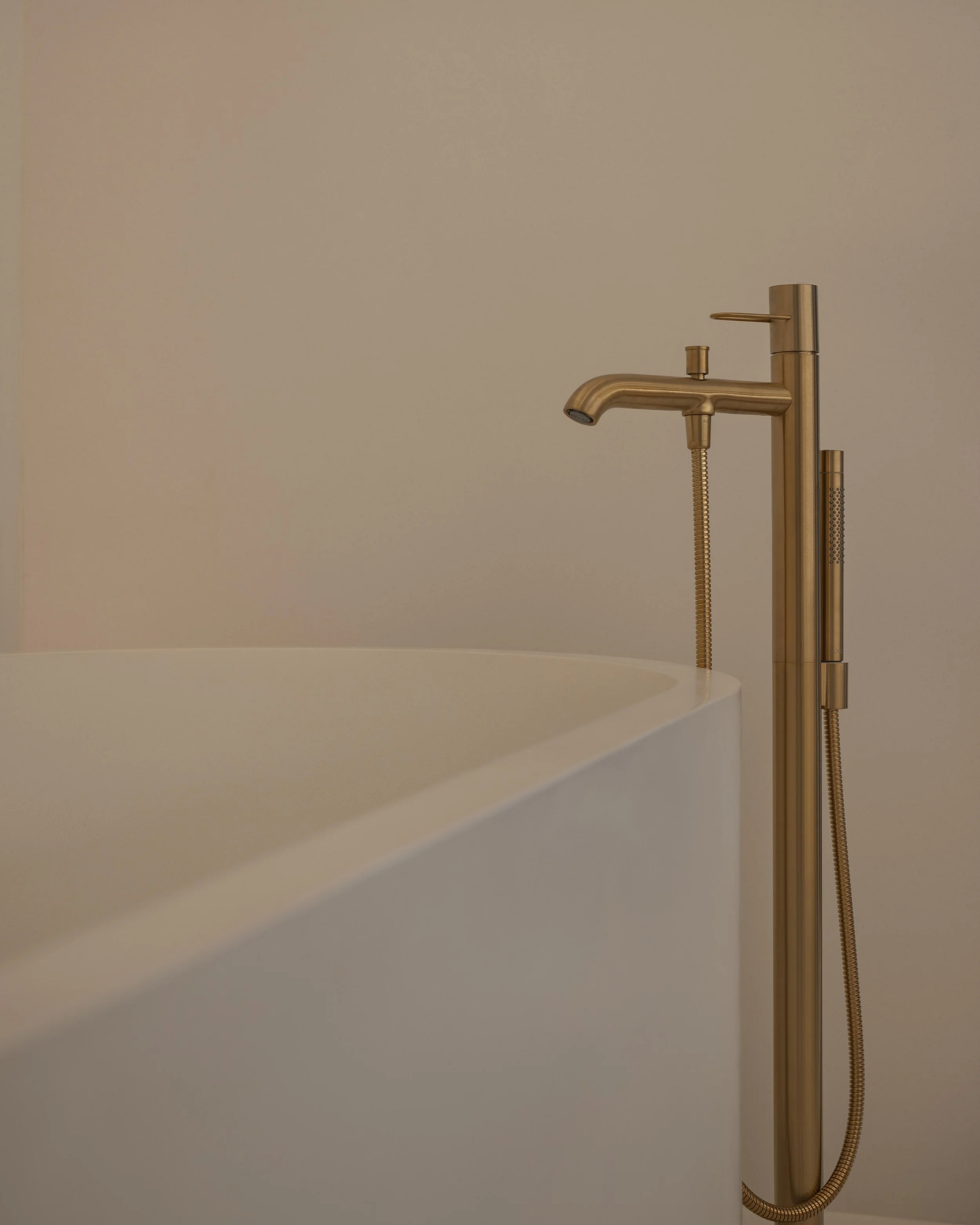 Close-up of a modern, gold-colored floor-mounted bathtub faucet with a handheld shower attachment against a beige wall.