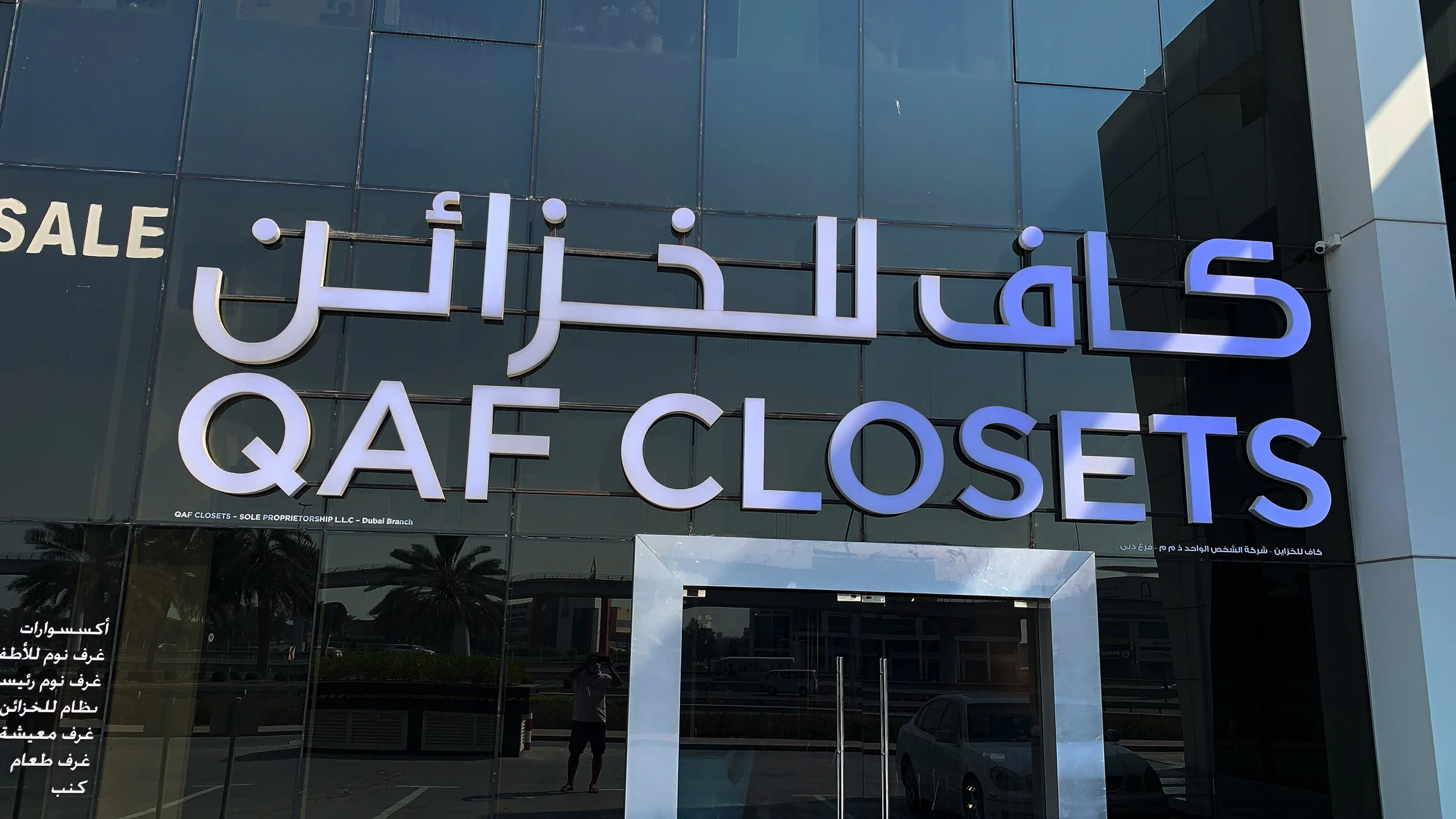 Sign for QAF Closets with Arabic writing above it, reflecting a palm tree and a person taking the photo in the glass