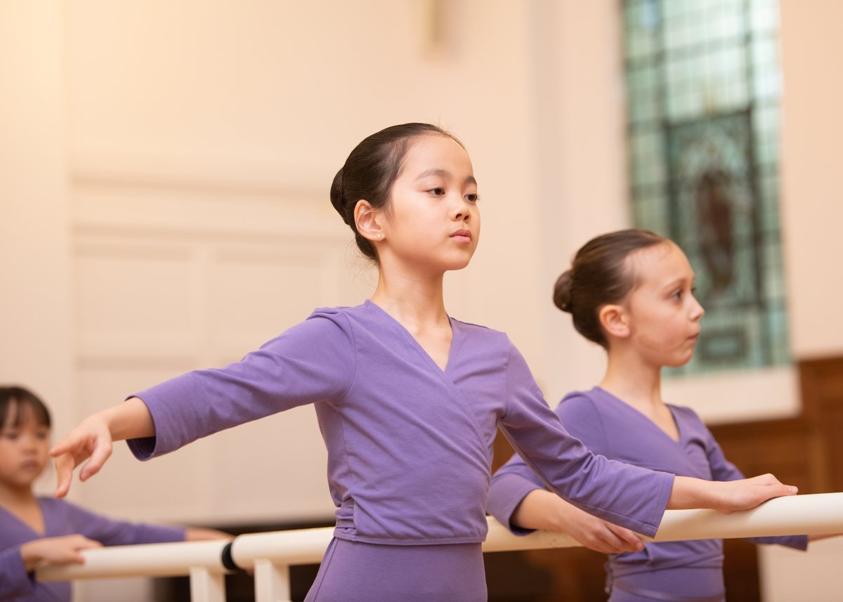 Young ballet dancers in purple uniforms practicing at a barre.