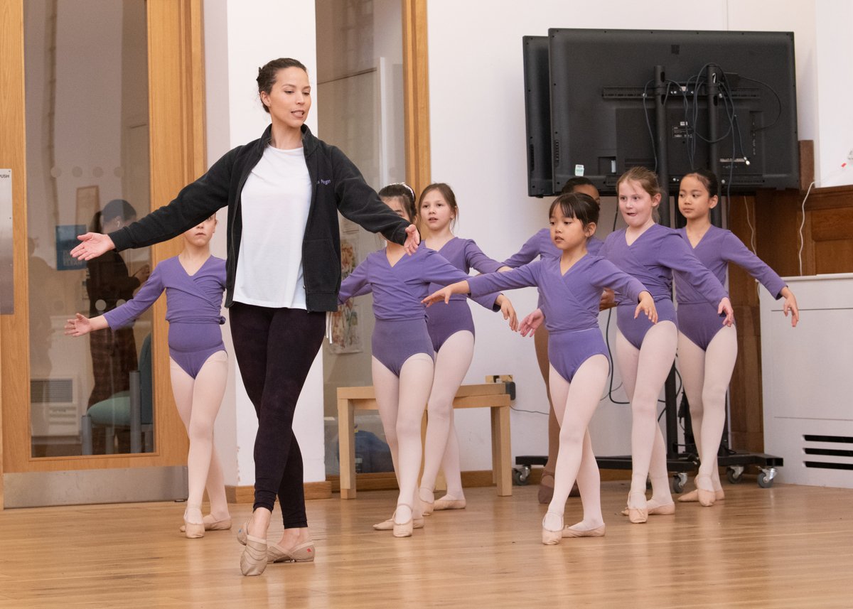 Ballet teacher instructing young girls in purple leotards during a dance class.