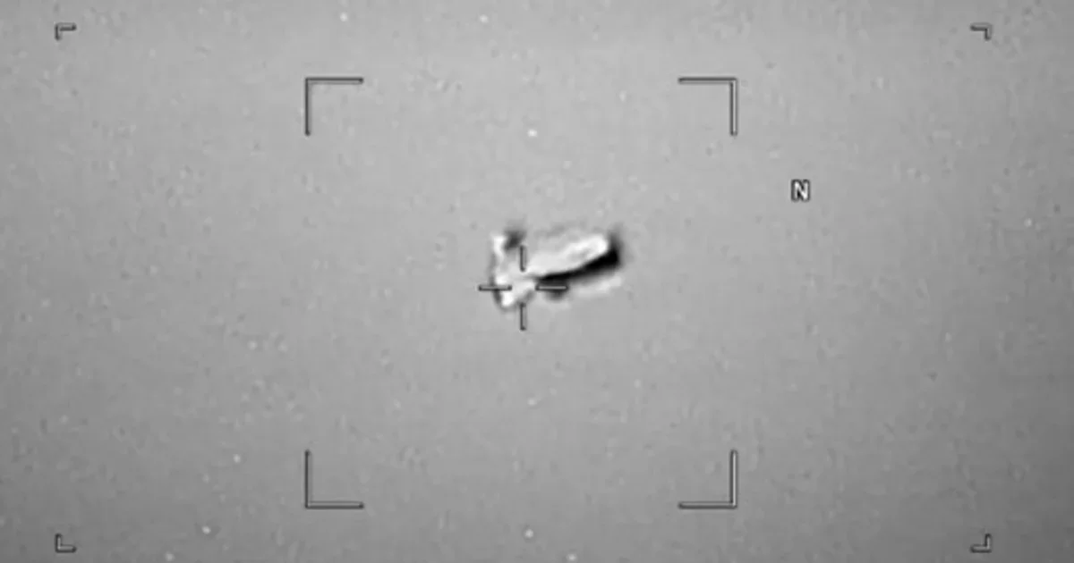 A black and white image taken through a targeting scope shows an unidentified flying object with a bright flash or explosion on its side, against a featureless, gray background.