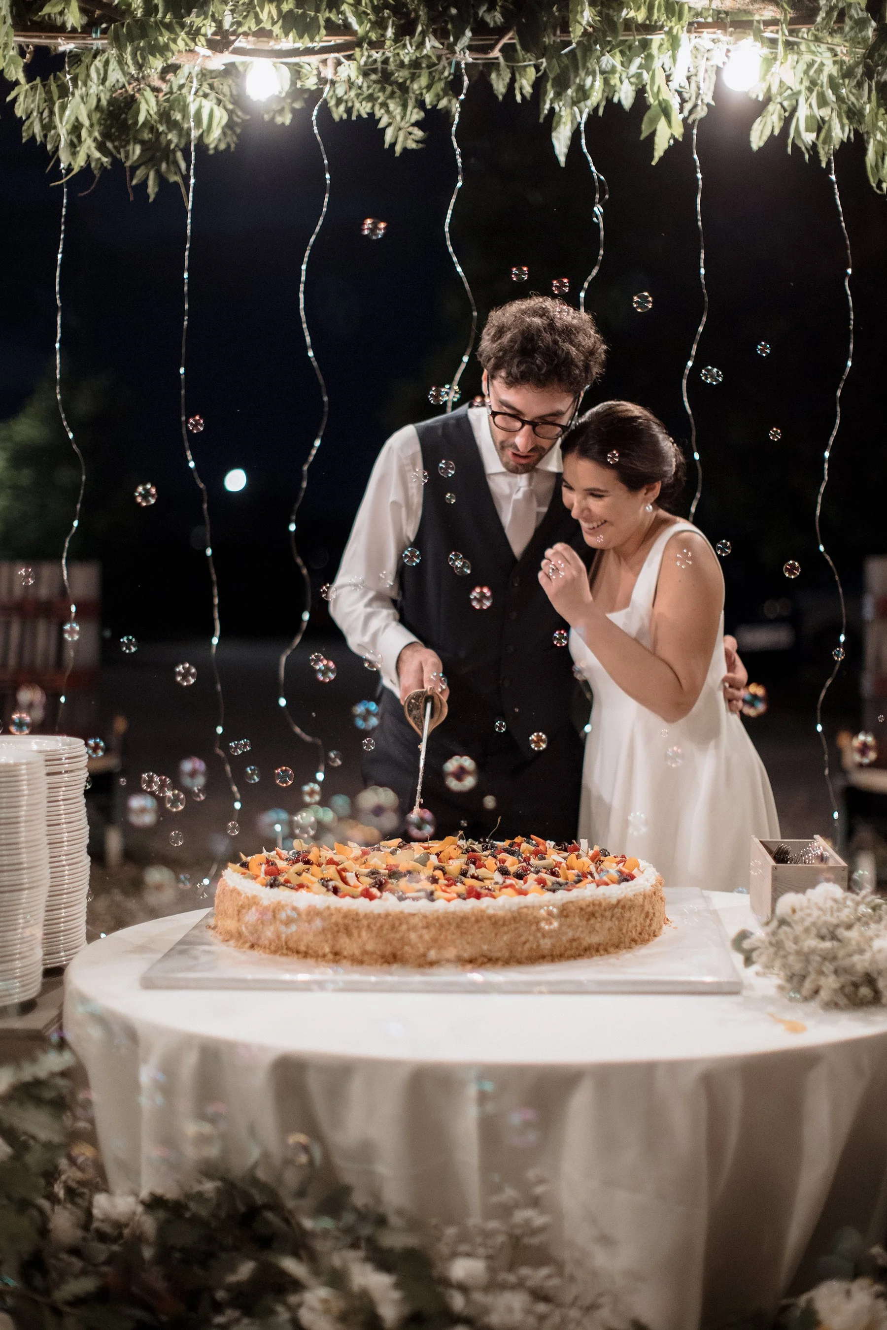 Coppia di sposi taglia una torta di matrimonio sotto luci decorative e fiori, con atmosfera serale romantica.