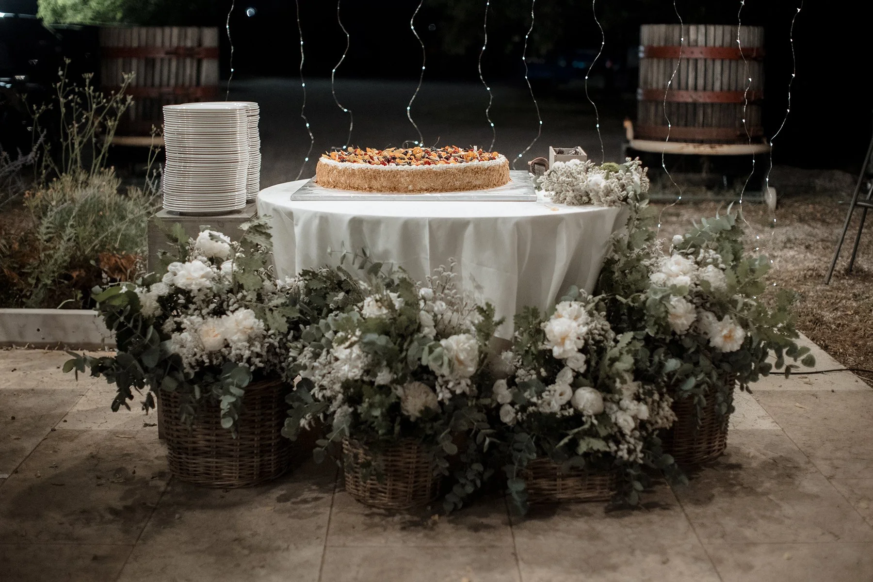 Tavolo decorato con un grande dolce circolare, fiori bianchi in vasi di vimini e ciotole di piatti bianchi, illuminato da luci stilo