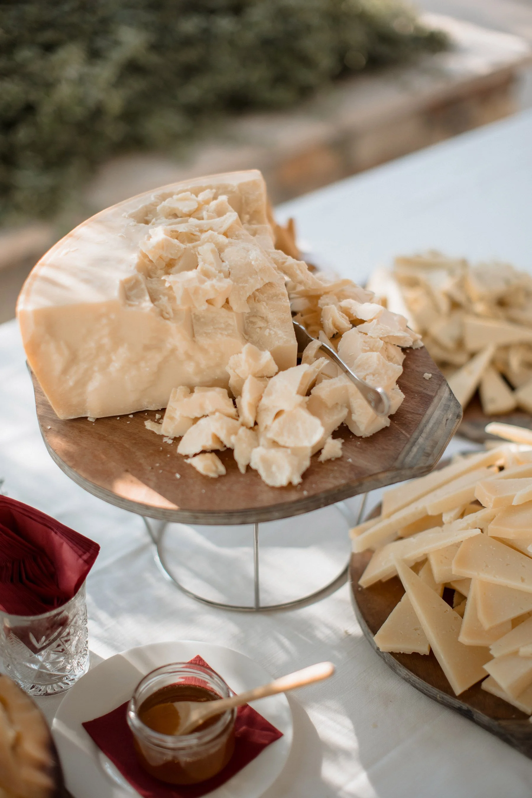 Formaggio a pasta dura e semi-dura messi su un vassoio di legno con coltelli, accompagnati da miele in un barattolo di vetro e coltello di legno, su tavolo bianco.