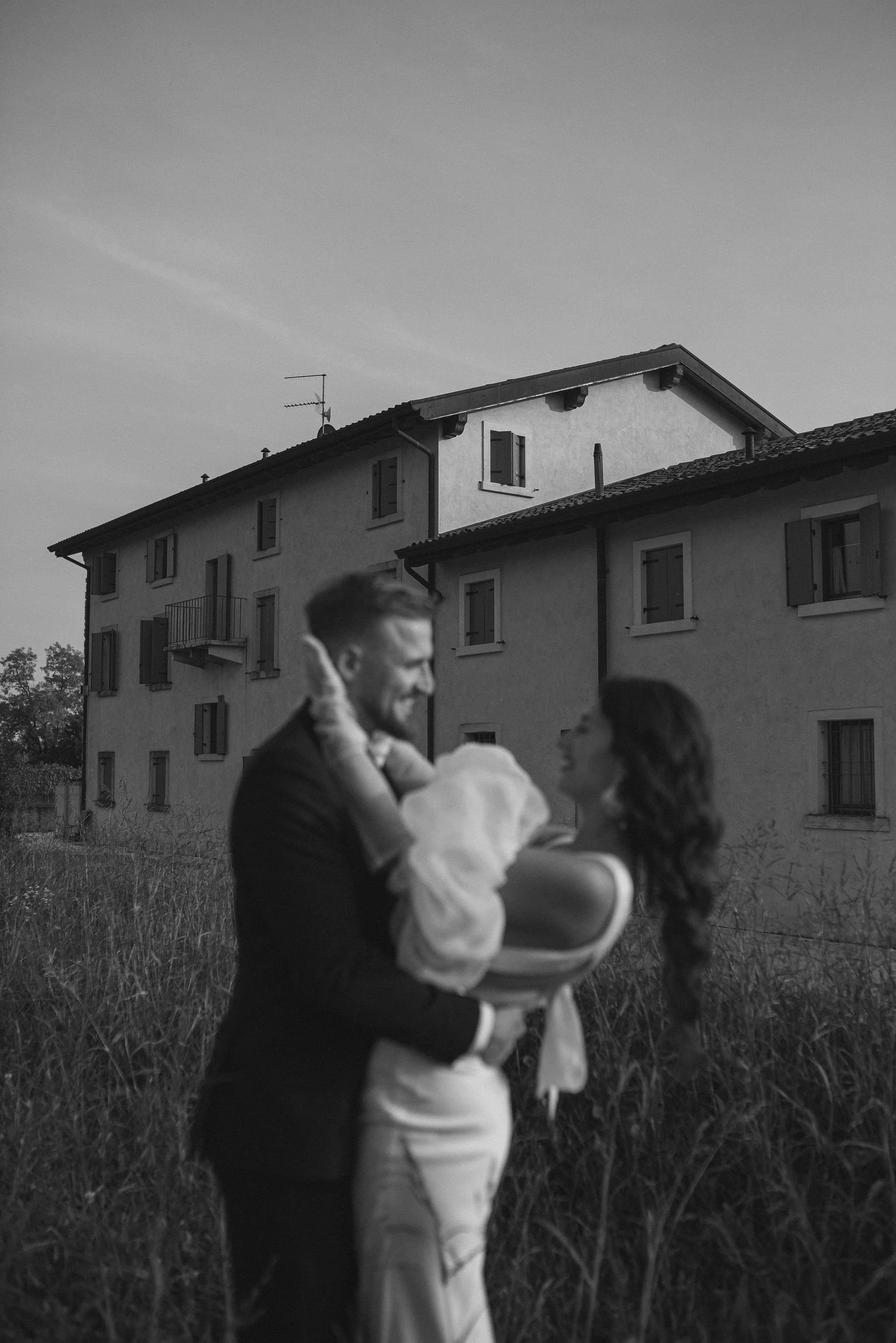 Una coppia con un bambino tra le braccia si abbracciano all'aperto davanti a un edificio con finestre e persiane durante il tramonto, in bianco e nero.