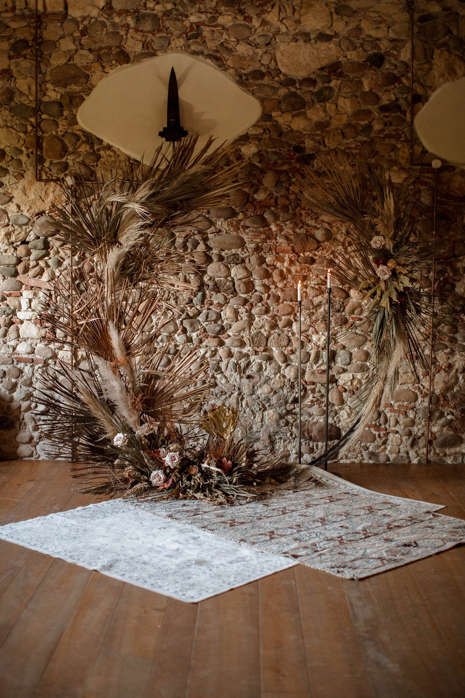 Decorazione floreale con rami secchi, fiori e piume disposta su un tappeto in una stanza con parete in pietra e luci soffuse.