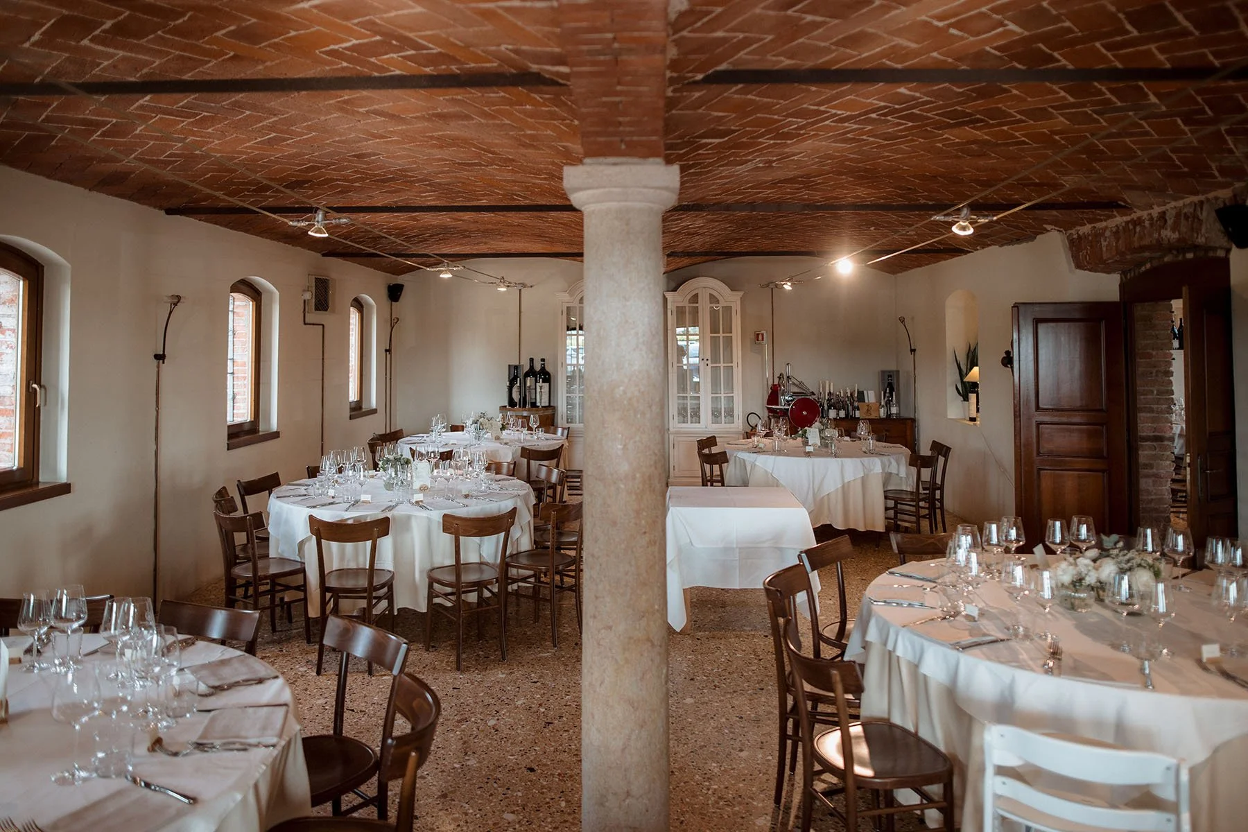 Sala da pranzo elegante con tavoli rotondi e rettangolari, apparecchiatura da cena con bicchieri e piatti, sedie in legno, pareti bianche, finestre con ante di legno, soffitto in mattoni con luci a incasso.