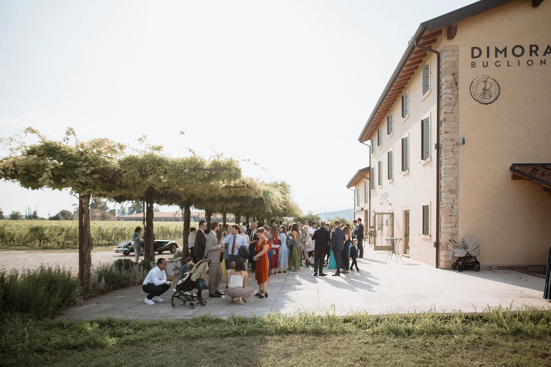 Gruppo di persone vestite elegantemente riunite all'aperto davanti a un edificio, con alcuni alberi e risaie sullo sfondo, durante una giornata di sole.