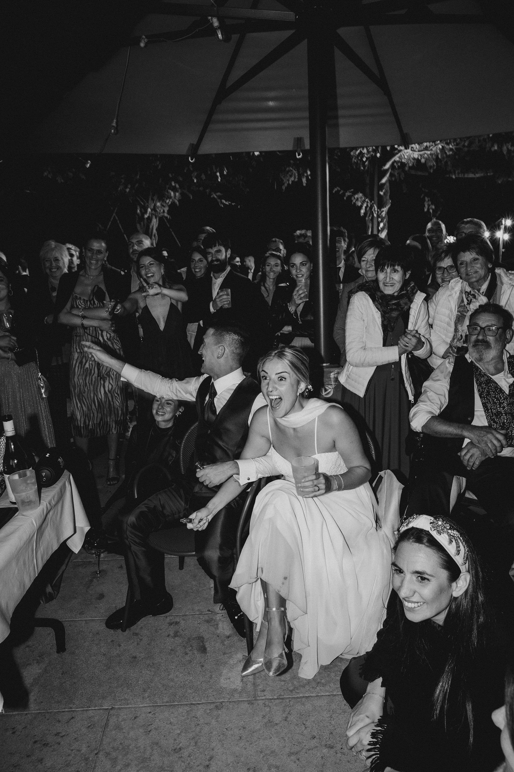 Numerose persone sorridenti e divertite a un evento di festa, con alcune sedute e altre in piedi, sotto un grande ombrello o tetto, illuminati dalla luce.