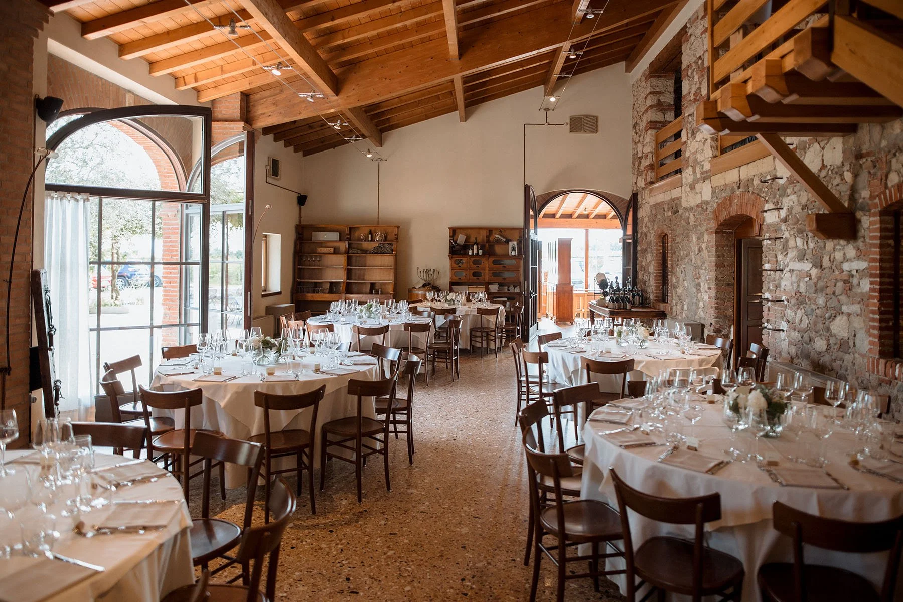 Sala per un evento con tavoli rotondi con tovaglie bianche, bicchieri e posate, sedie di legno, pareti in pietra e mattoni, soffitto in legno con travi, grande porta affacciata su un giardino e scaffali di legno con decorazioni.