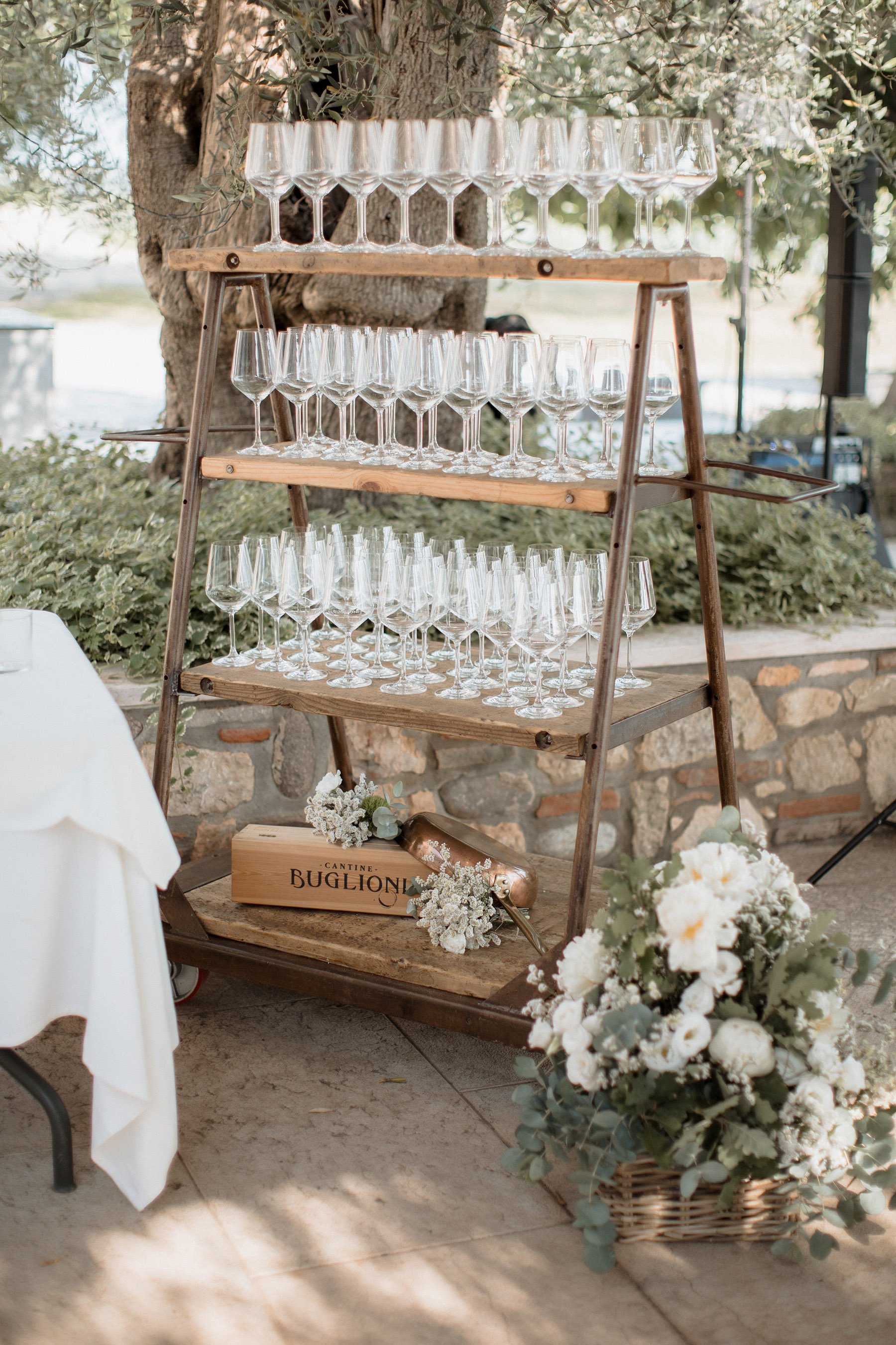 Vassoio con bicchieri da vino su scaffale in legno, decorato con fiori bianchi e un cartellino con scritto 'Cantine Buglioni', all'aperto con alberi e vegetazione.