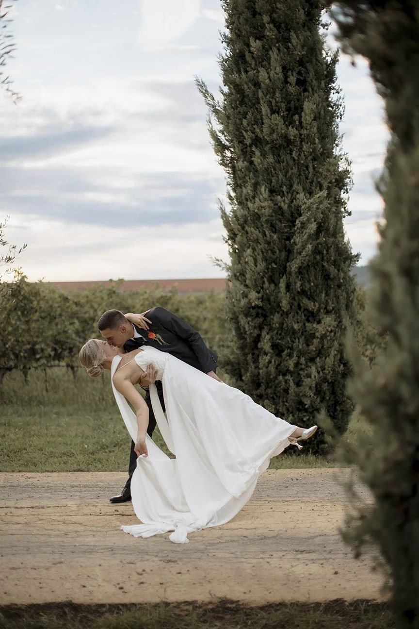 Un uomo e una donna in abiti da matrimonio ballano all'aperto, lui sta dipingendo la donna all'indietro, circondati da alberi con il cielo nuvoloso sullo sfondo.