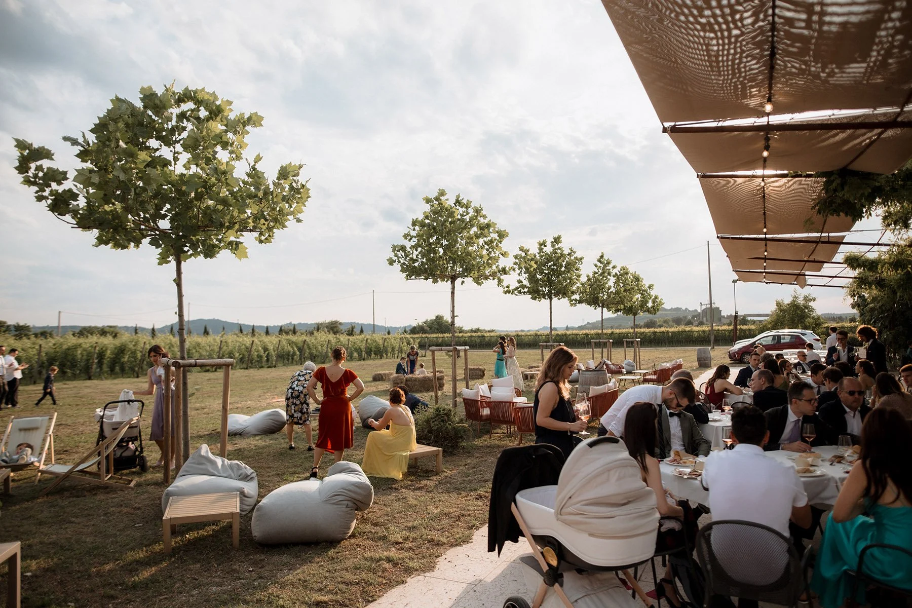 Festeggiamento all'aperto con persone sedute a tavoli, donne in abiti eleganti, bambini, un'area con alberi e un campo verde, sotto un'ampia tenda.