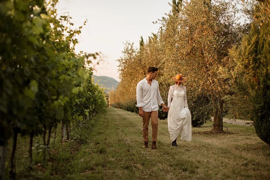 Una coppia cammina mano nella mano tra filari di viti in un vigneto, in una giornata di sole, con uomini e donne vestiti elegantemente, la donna indossa un vestito bianco e un cappello a tesa larga.