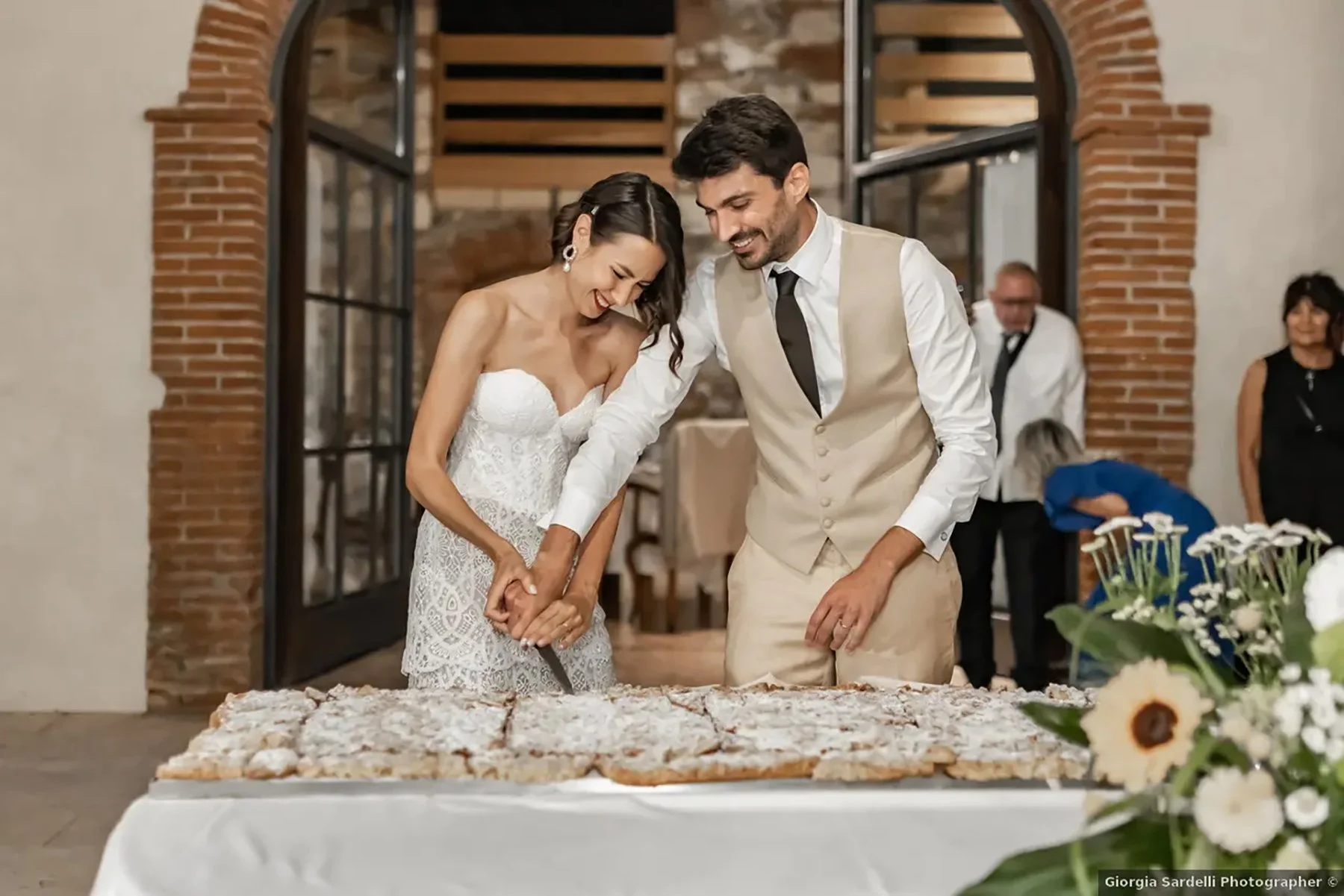 Una coppia di sposi taglia insieme una torta di nozze durante un ricevimento di matrimonio in un locale con pareti di mattoni e decorazioni floreali.